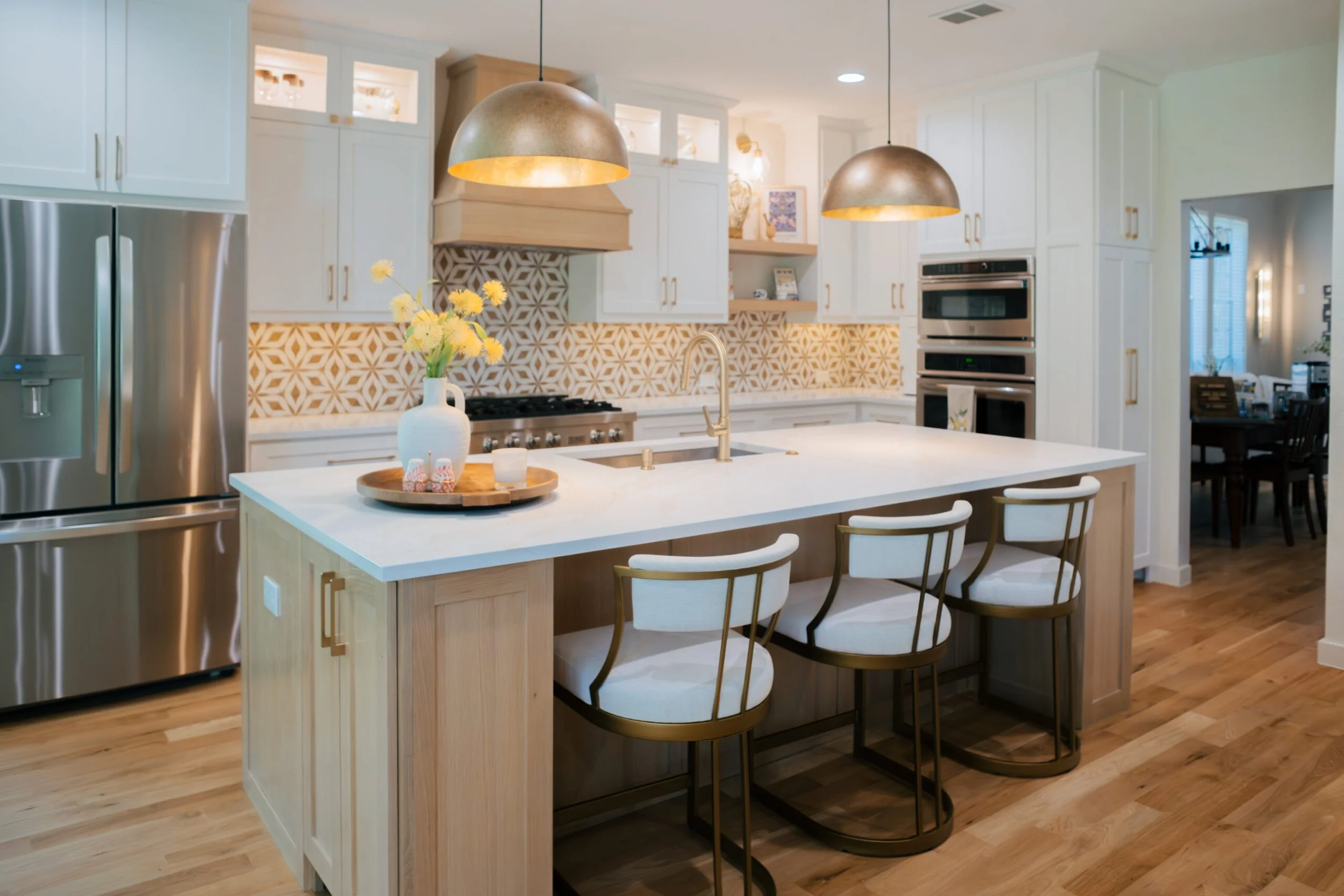 Modern kitchen with white cabinets, a white island, gold hardware, pendant lighting, a floral vase on the island, and geometric patterned backsplash.