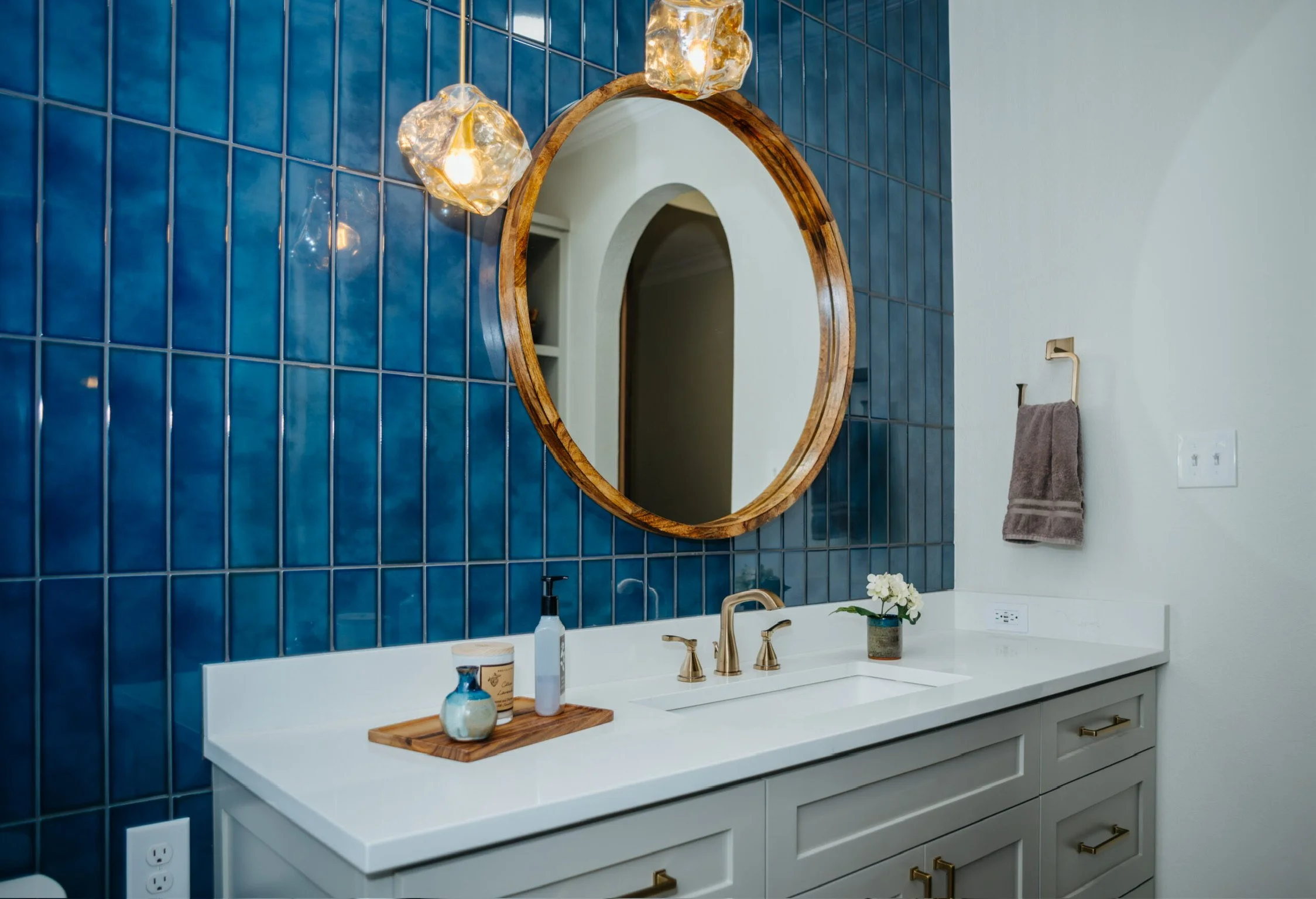 A modern bathroom vanity area featuring a large round wooden-framed mirror mounted on a blue tiled wall, with two hanging glass pendant lights, a white countertop with a gold faucet, and various decor items including small plants and toiletries.