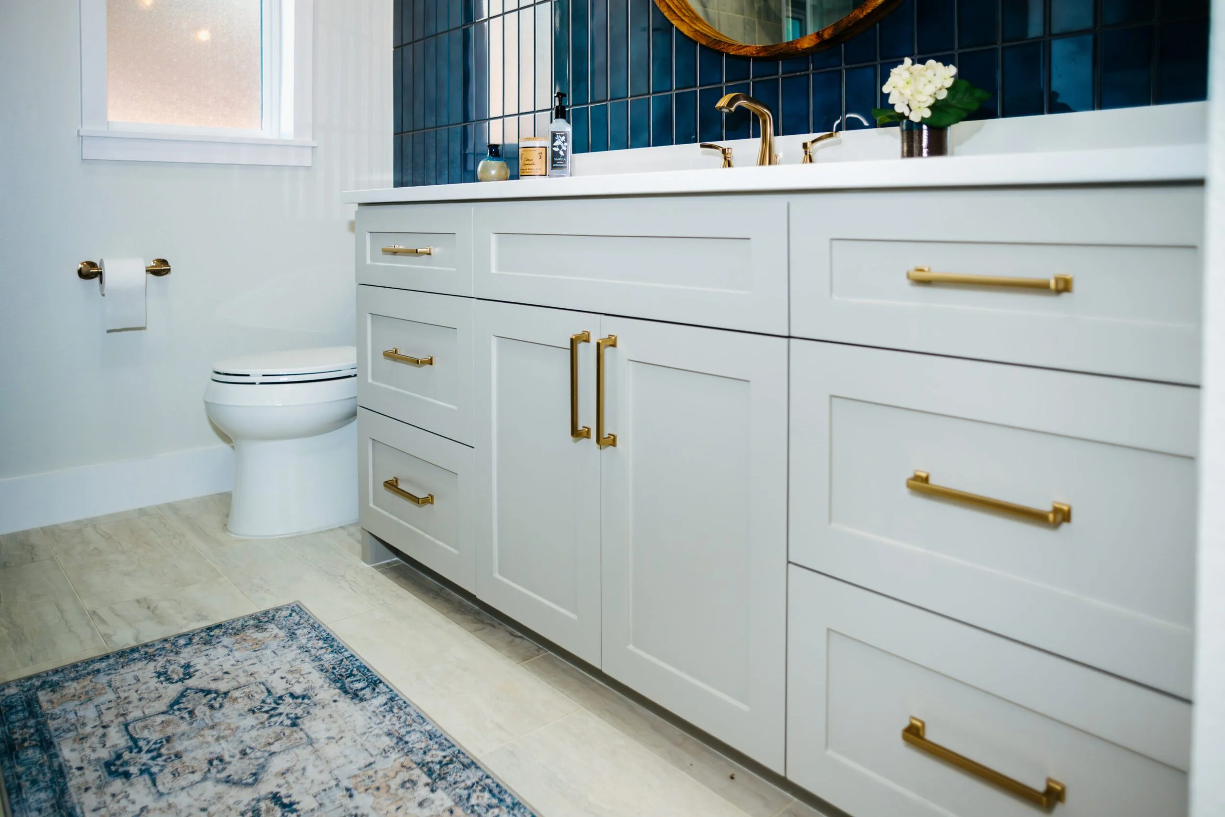 A modern bathroom with a white vanity cabinet featuring gold handles, a marble countertop, and a gold faucet. There is a blue tiled wall behind the vanity, a mirror above it, a potted white hydrangea flower, and various toiletries. The bathroom has a