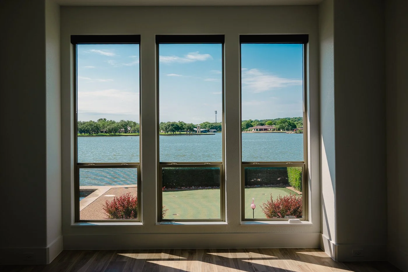 View of a lake seen through a large three-pane window in a room, with a grassy yard, shrubs, and trees across the water under a partly cloudy sky.