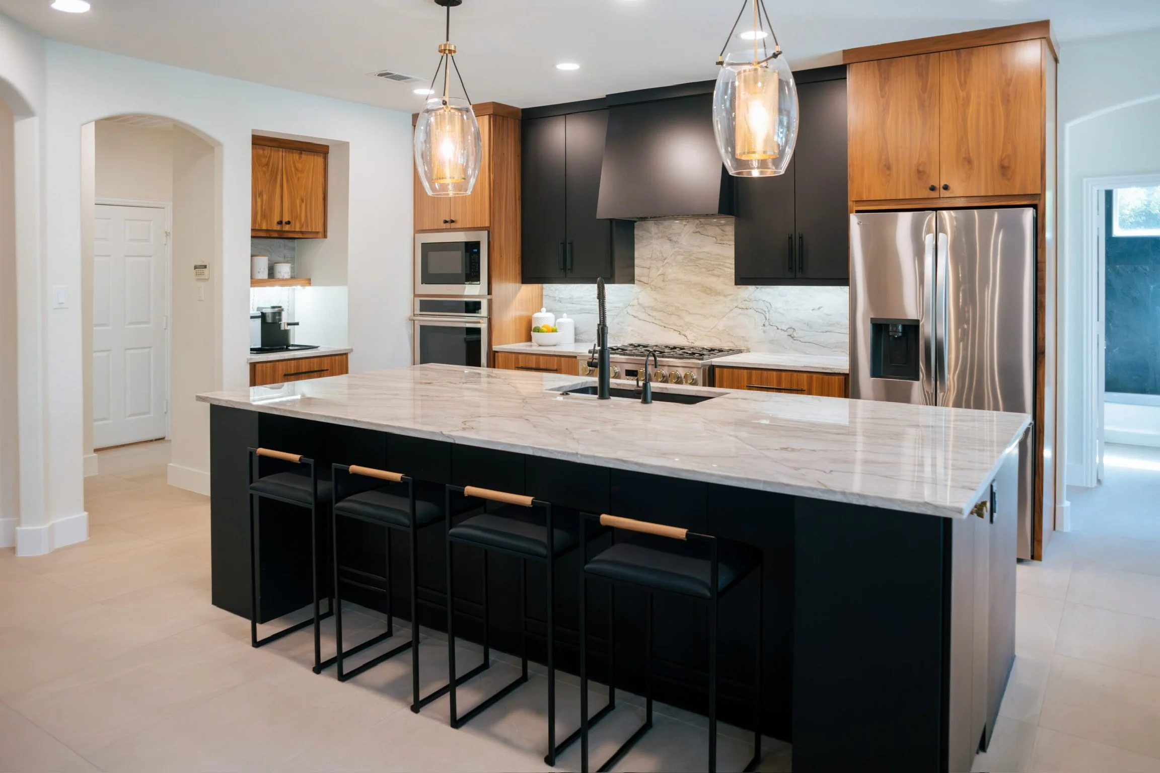 Modern kitchen with black and wood cabinetry, marble backsplash, white tile floors, large island with a marble countertop, black bar stools, and gold-accented pendant lighting.