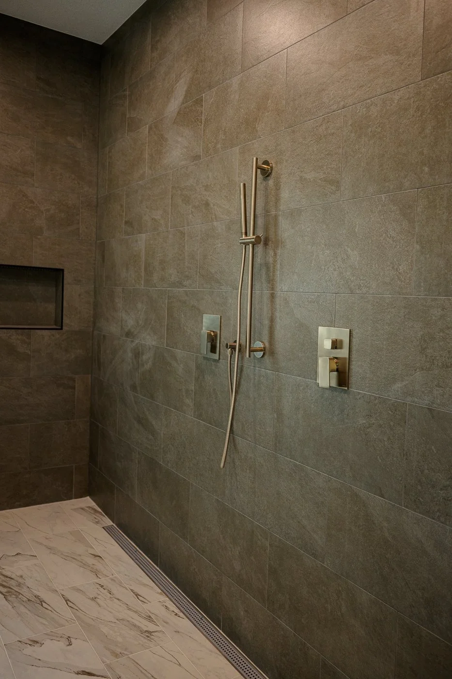 Modern shower with gray stone tile wall, handheld showerhead, and two gold fixtures, marble floor, and built-in niche.