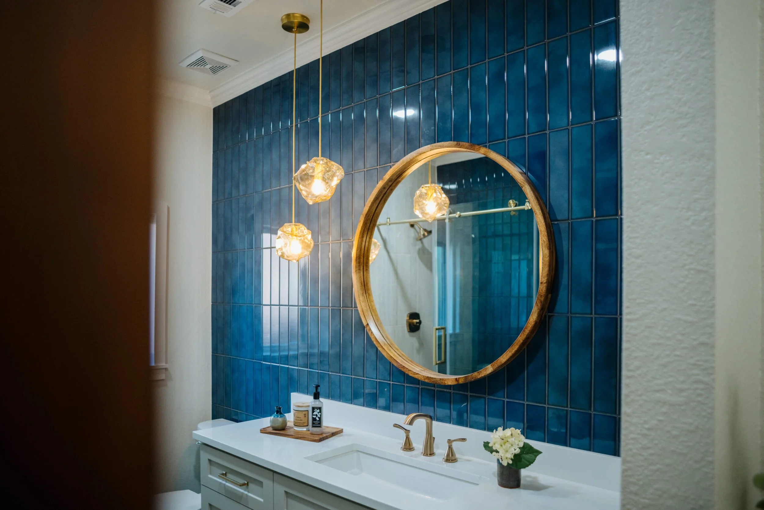 Bathroom with a white vanity, gold fixtures, and a blue tiled wall. A round wooden mirror hangs above the sink, decorated with potted flowers and decor.