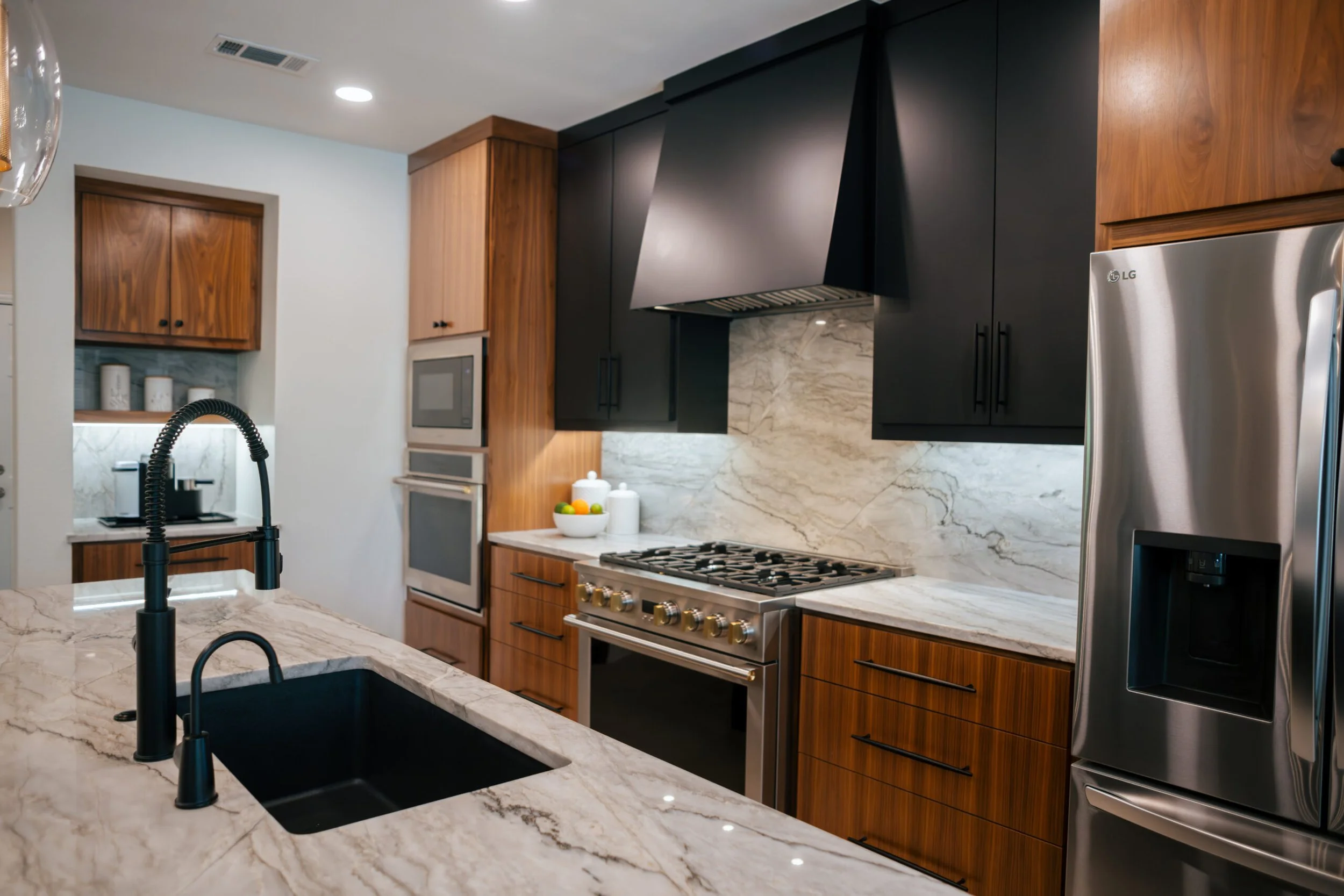 Modern kitchen with black and wooden cabinets, marble countertops, stainless steel appliances, black sink with a black faucet, and decorative jars on the countertop.