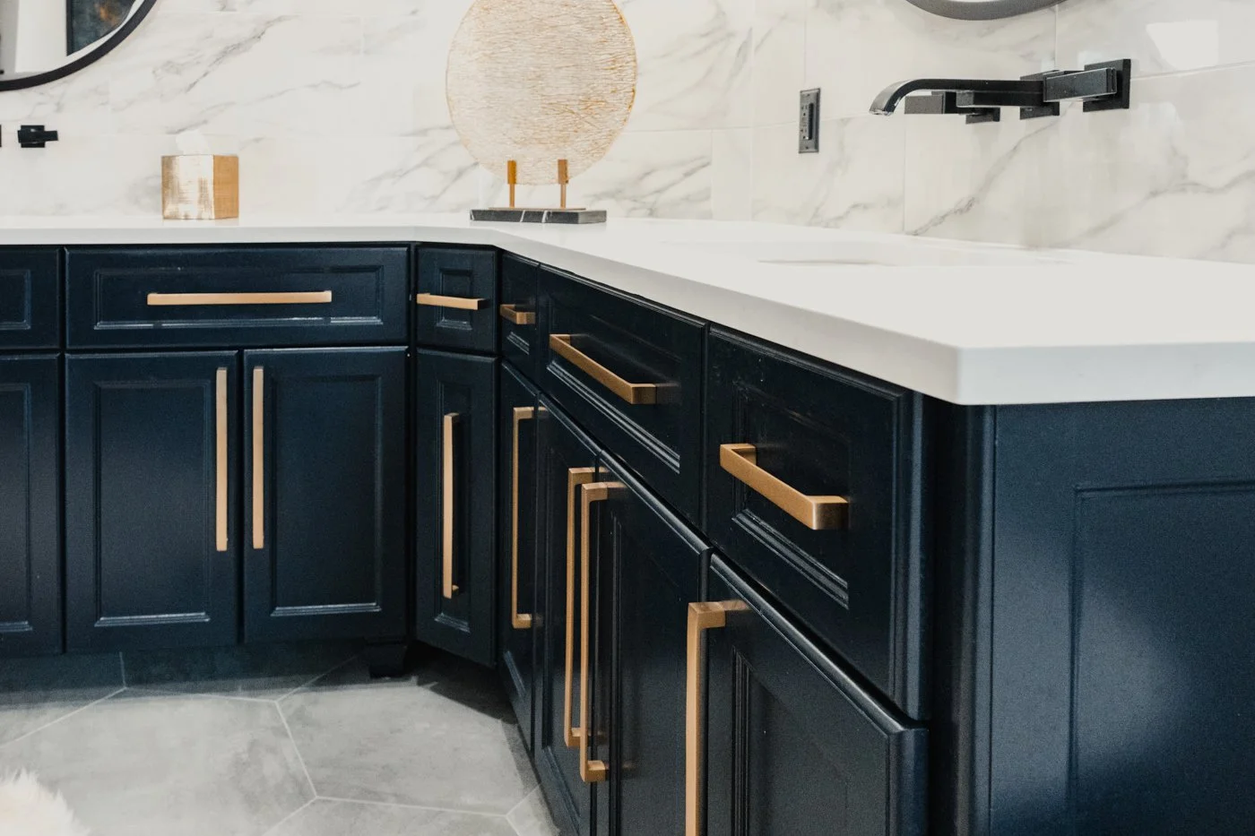 Close-up of a modern kitchen countertop with dark blue cabinets featuring gold handles, a white stone countertop, and a marble backsplash with a circular decor item.