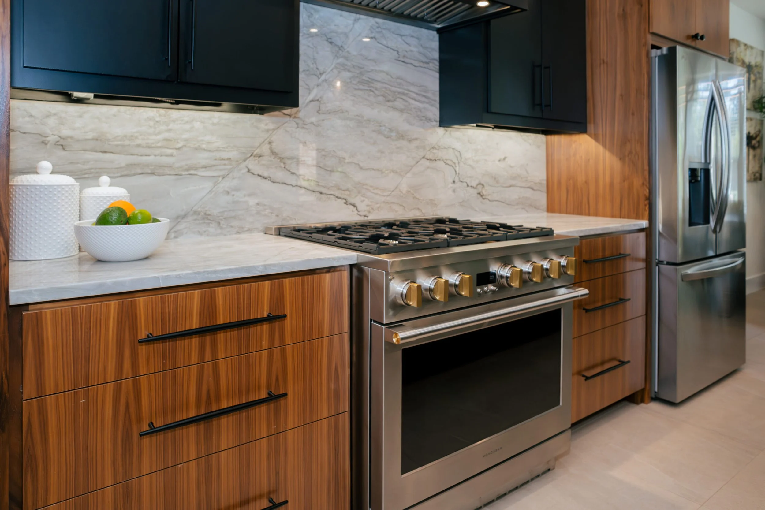 Modern kitchen with wood cabinets, marble countertops, and a stainless steel gas stove. There are two white jars, a bowl of limes and oranges, and a stainless steel refrigerator.