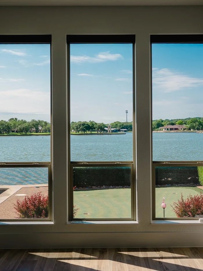 View through three large windows showing a lake, green trees, a building, a radio tower, and a partly cloudy sky.