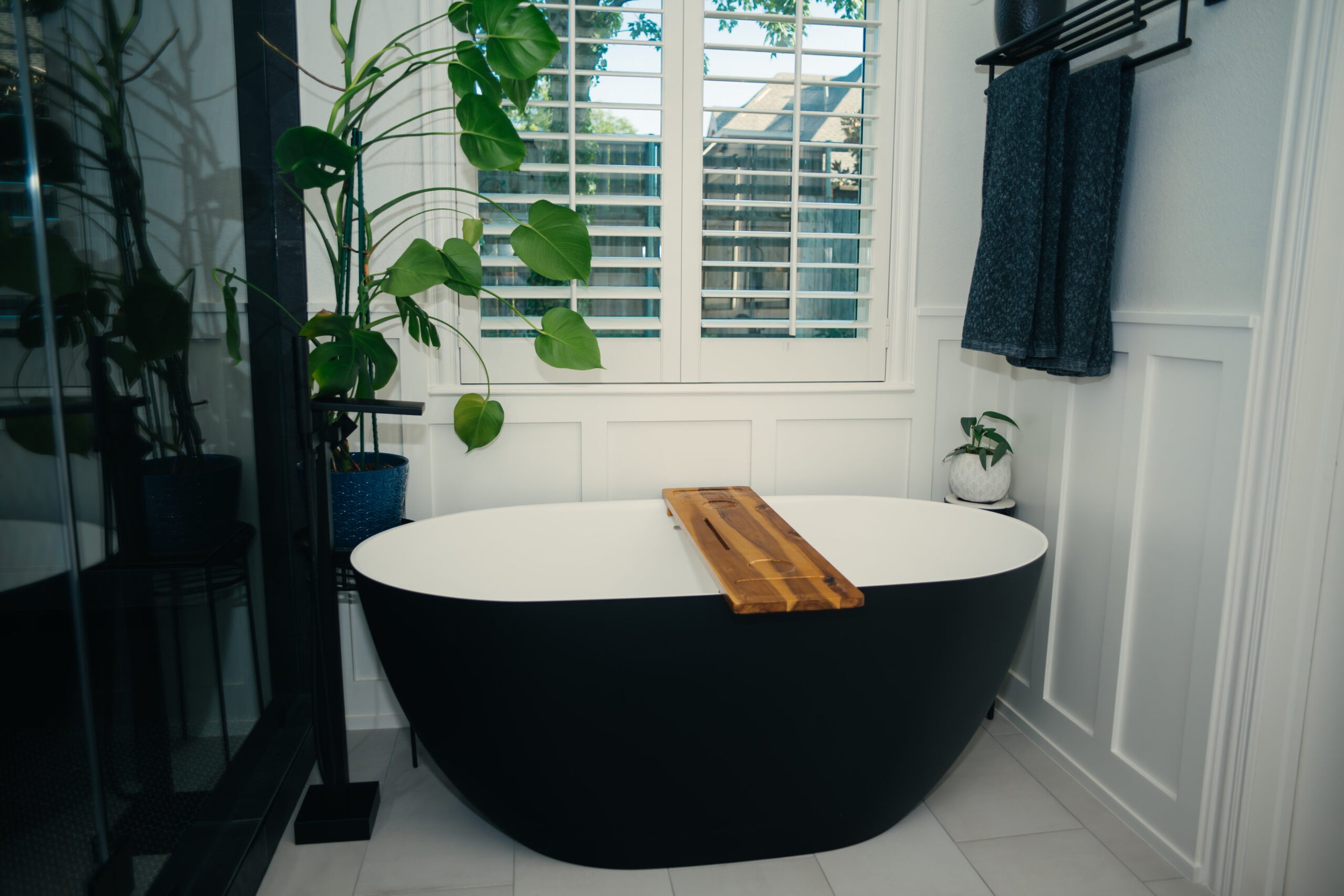 A modern bathroom with a black bathtub with white interior and wooden tray, window with white shutters, green houseplants, towel rack with dark towels, and white paneled walls.