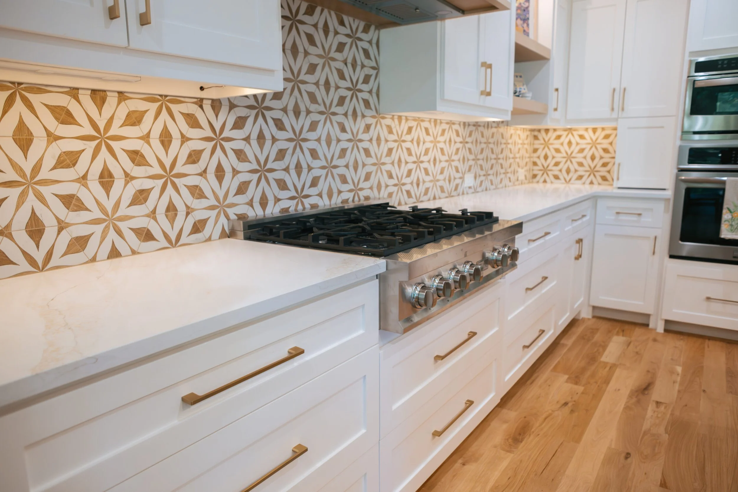 Kitchen with white cabinets, gold handles, patterned tile backsplash, gas stove, and hardwood flooring.