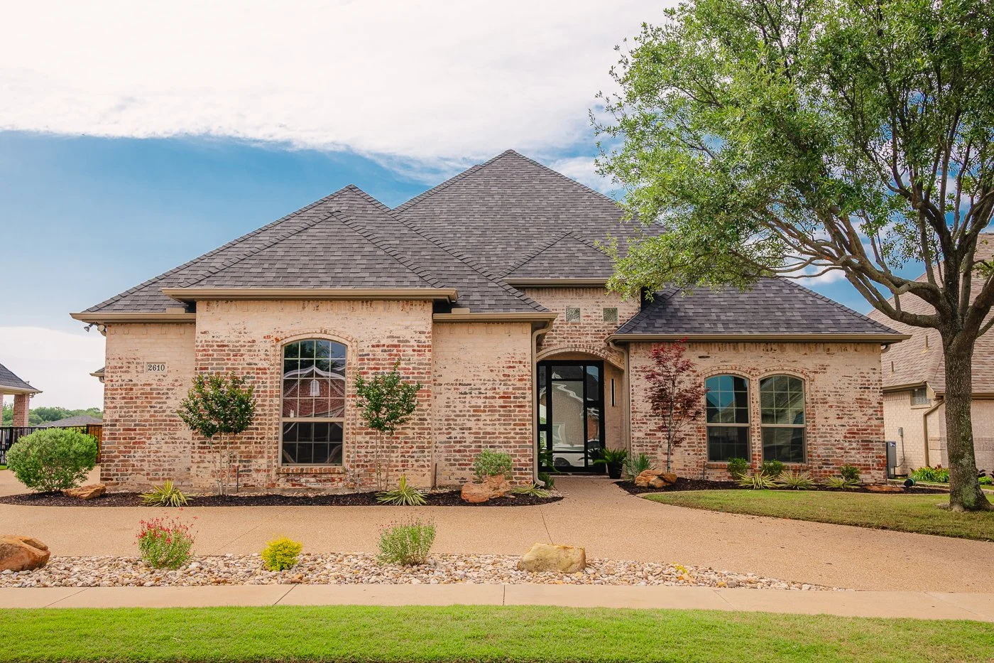 Front view of a brick house with a driveway, green lawn, trees, and plants, under a partly cloudy sky.