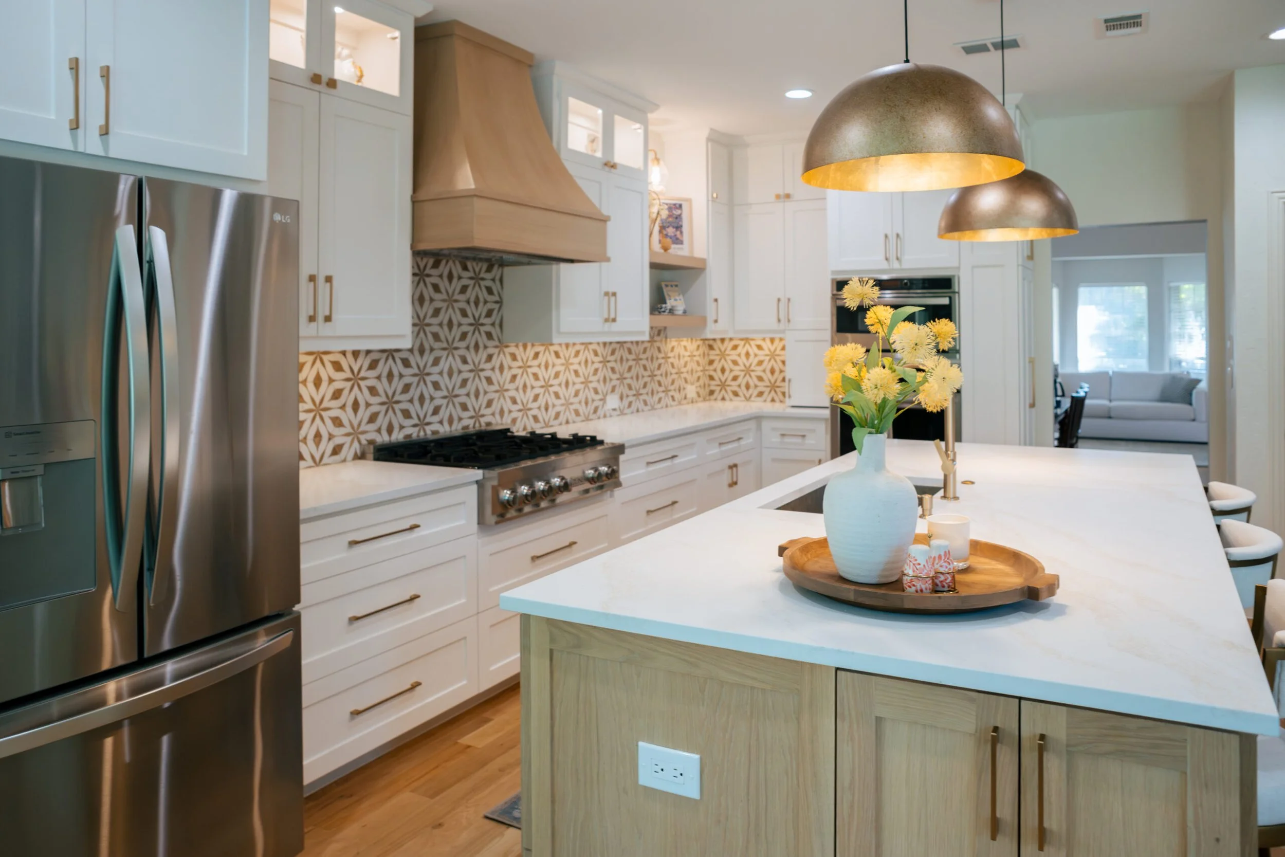 Modern kitchen with white cabinets, a stainless steel fridge, a wooden range hood, patterned backsplash, a white kitchen island with a vase of yellow flowers and a tray of small gifts, and gold pendant lights hanging above.