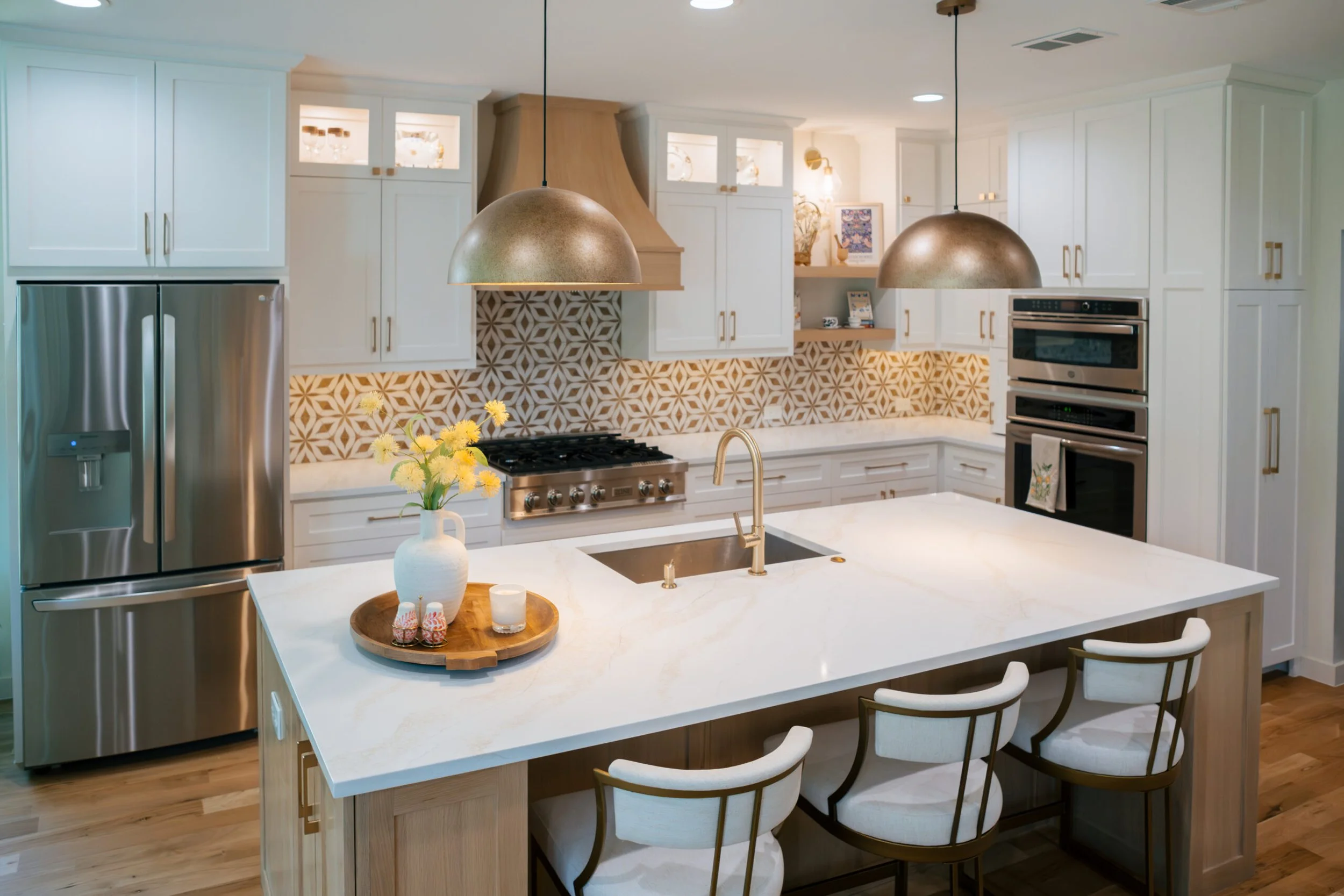 Modern kitchen with white cabinetry, patterned backsplash, and large island. Pendant lights hang over the island, which has a vase with yellow flowers and a tray with small items. Stainless steel refrigerator and oven are visible.