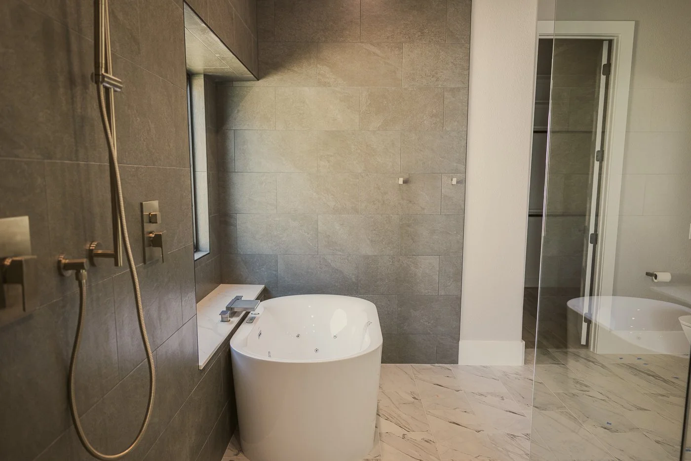 Modern bathroom with a white bathtub, gray tiles on the wall, a glass shower door, and a marble floor.