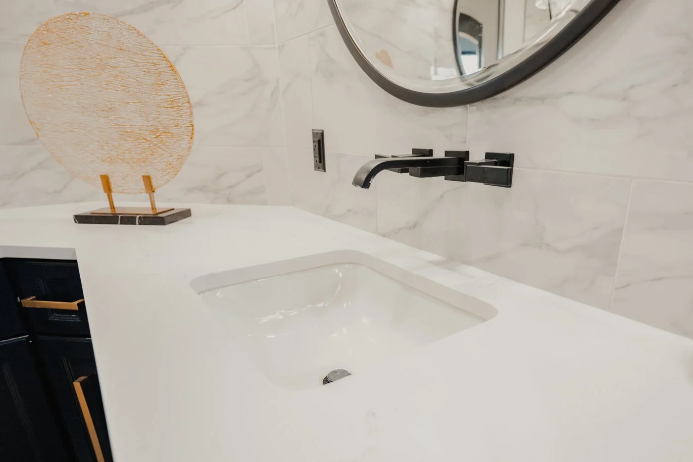 Modern bathroom with white marble backsplash, black faucet, round mirror, and a decorative orange and gold sculpture on the counter.
