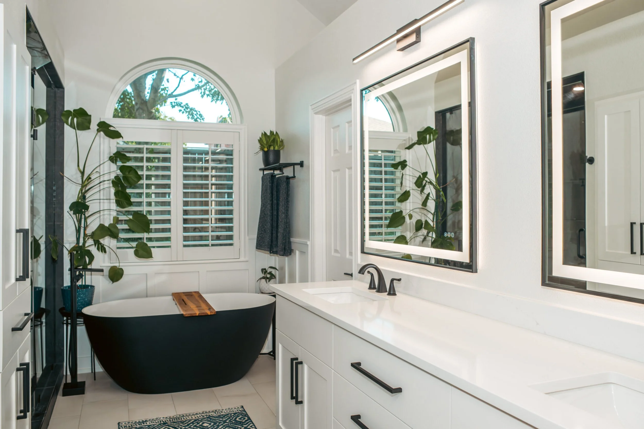 Modern bathroom with white walls and cabinetry, black bathtub, large window with plantation shutters, and several green plants.