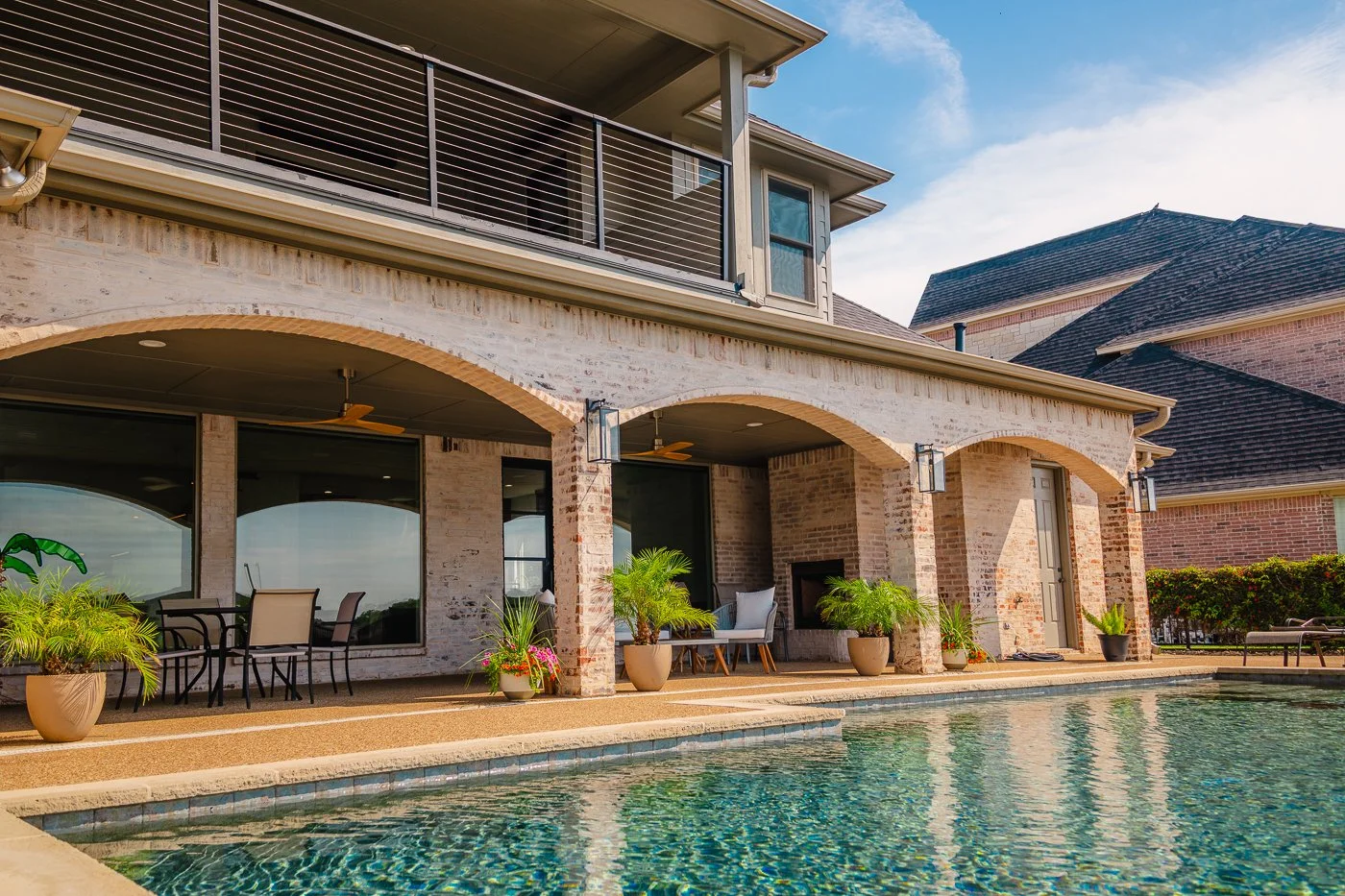 Backyard with swimming pool, patio seating, potted plants, and a two-story house with brick and siding exterior and large windows.
