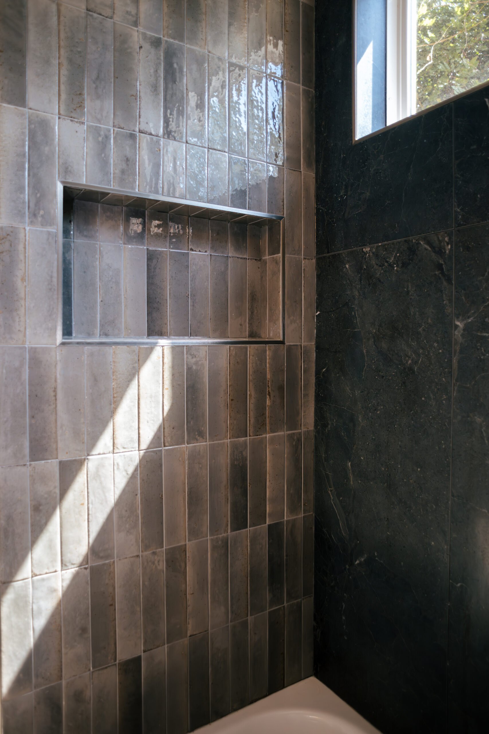 Bathroom with beige tiled shower area, built-in shelf, black marble wall, and window with trees outside.