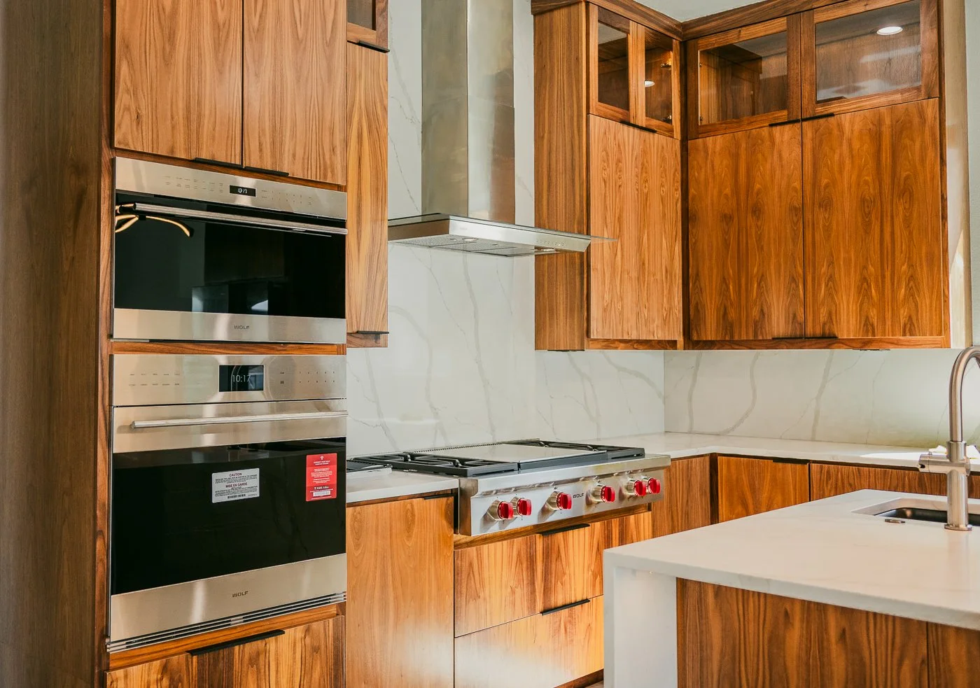 Modern kitchen with wooden cabinets, stainless steel oven, stovetop with red control knobs, marble backsplash, and a white countertop with sink.