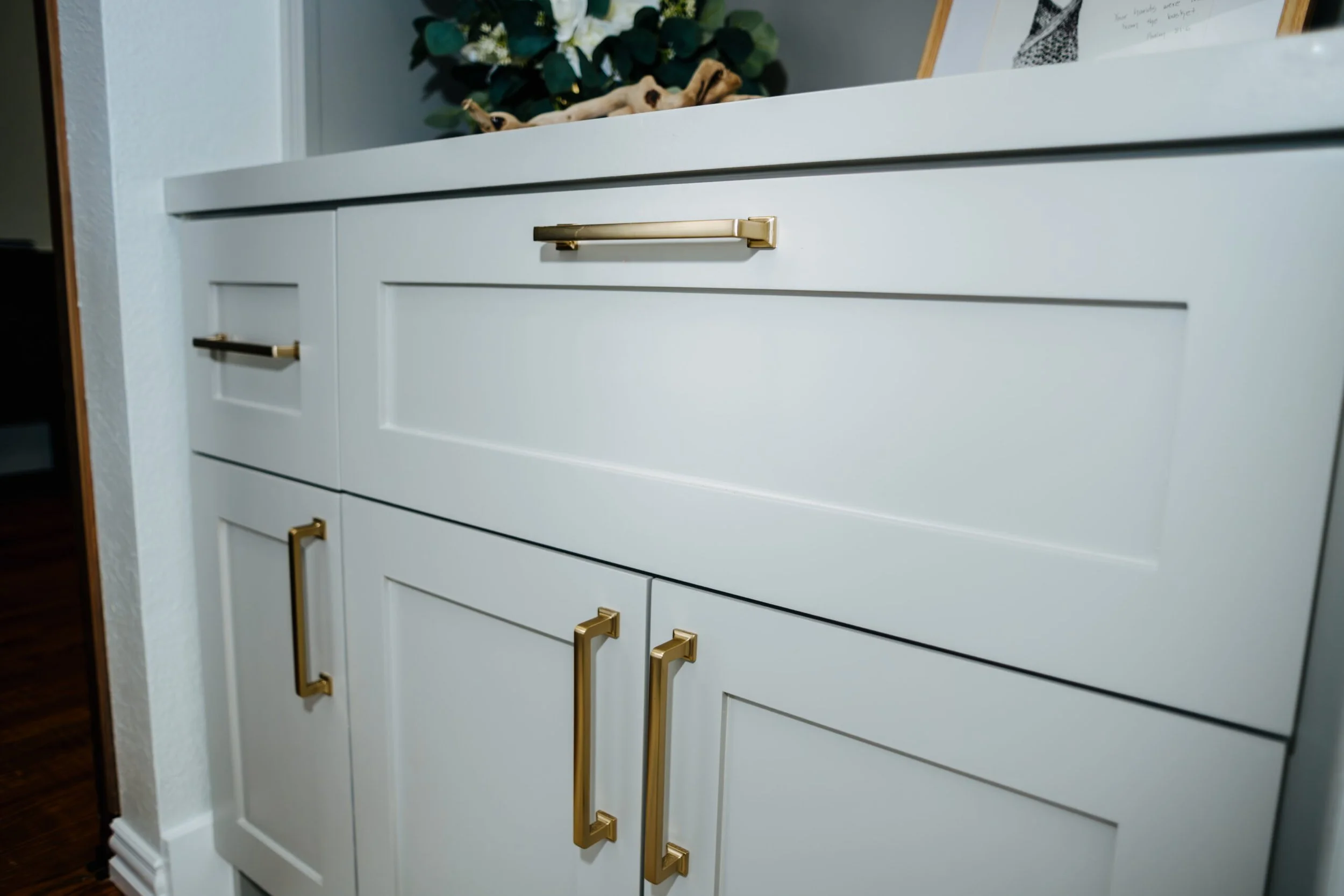 A white dresser with five drawers and gold handles, positioned against a wall with some decorative items on top, including a framed photograph or artwork, a floral arrangement with white flowers, and a twisted wooden sculpture.