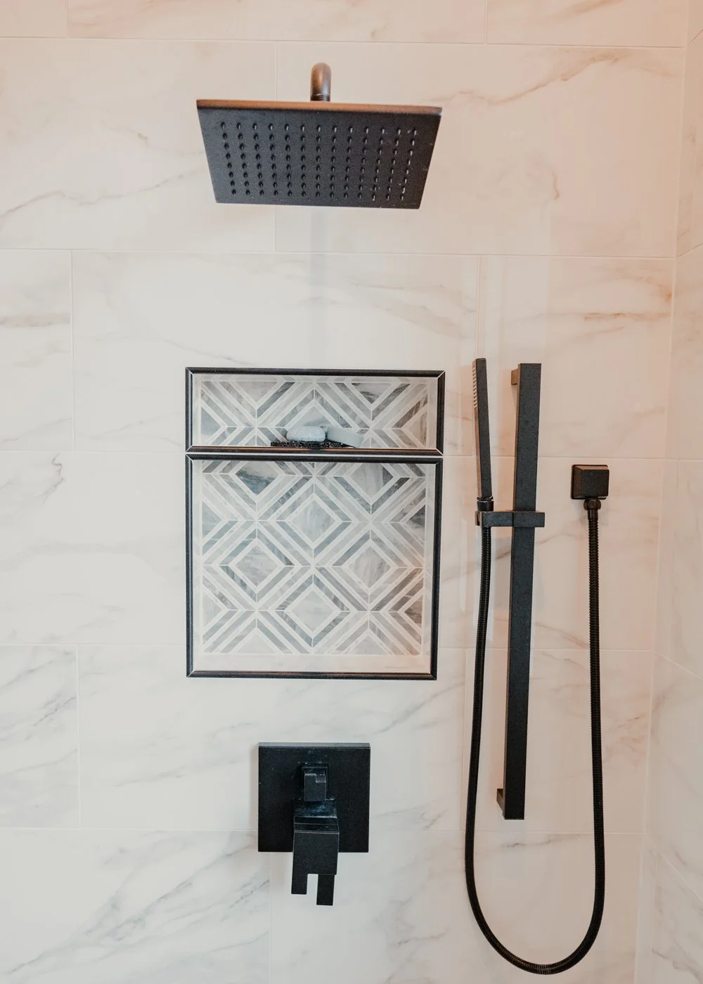 Modern walk-in shower with black square rain showerhead, a textured soap dish, and a black handheld shower wand mounted on marble walls.