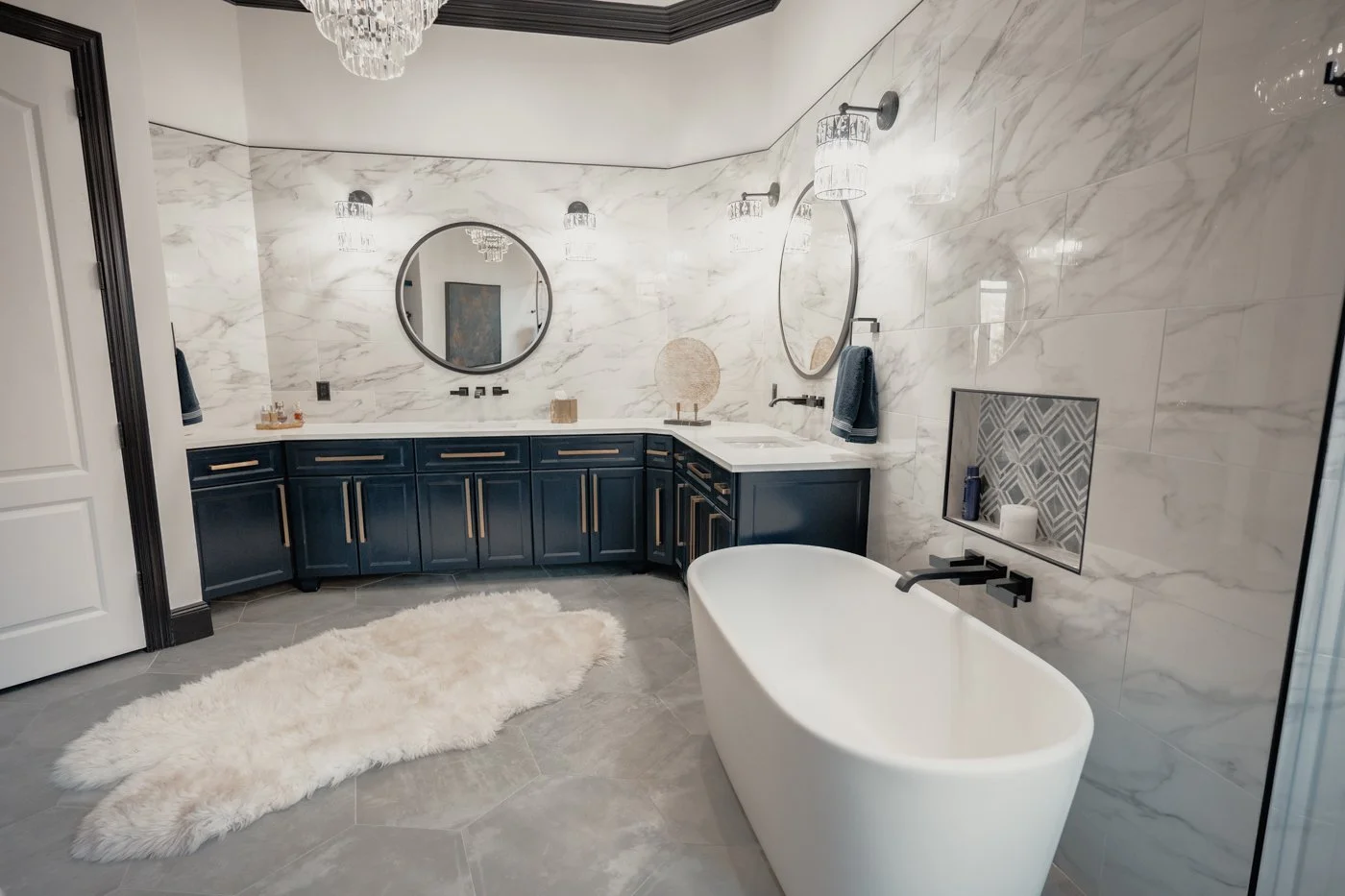 Modern bathroom with marble walls, dark blue cabinets with gold handles, a white freestanding bathtub, two large round mirrors above the vanity, and a fluffy white rug on grey tile flooring.