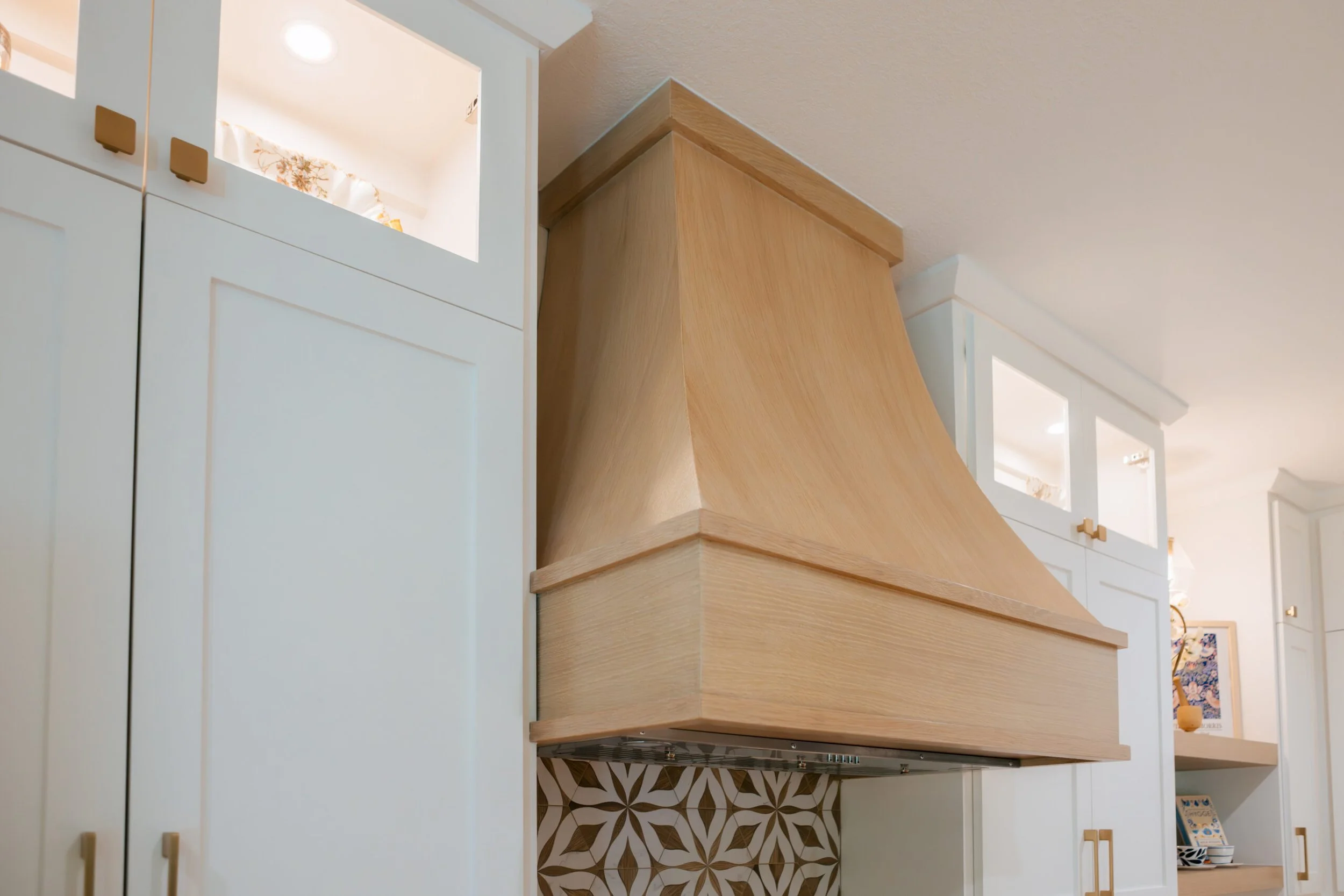 Close-up of a modern kitchen with white cabinets, gold handles, a wooden range hood, and decorative patterned tiles on the wall.