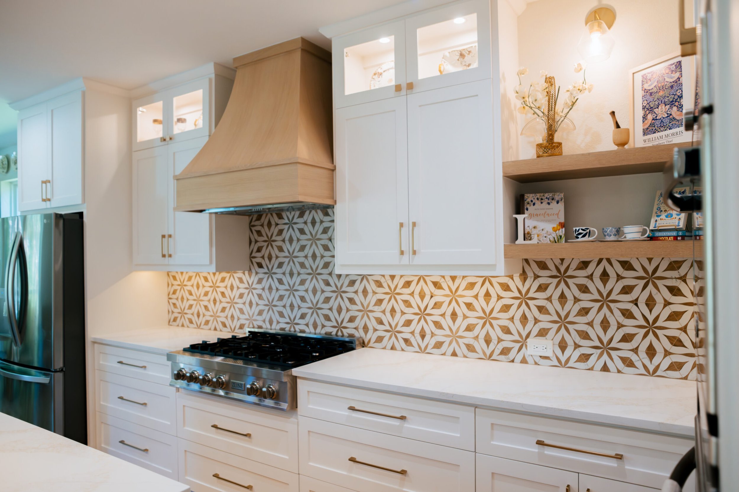 Modern kitchen with white cabinets, a patterned tile backsplash, a wooden range hood, and open shelves displaying books, mugs, and decorative items.