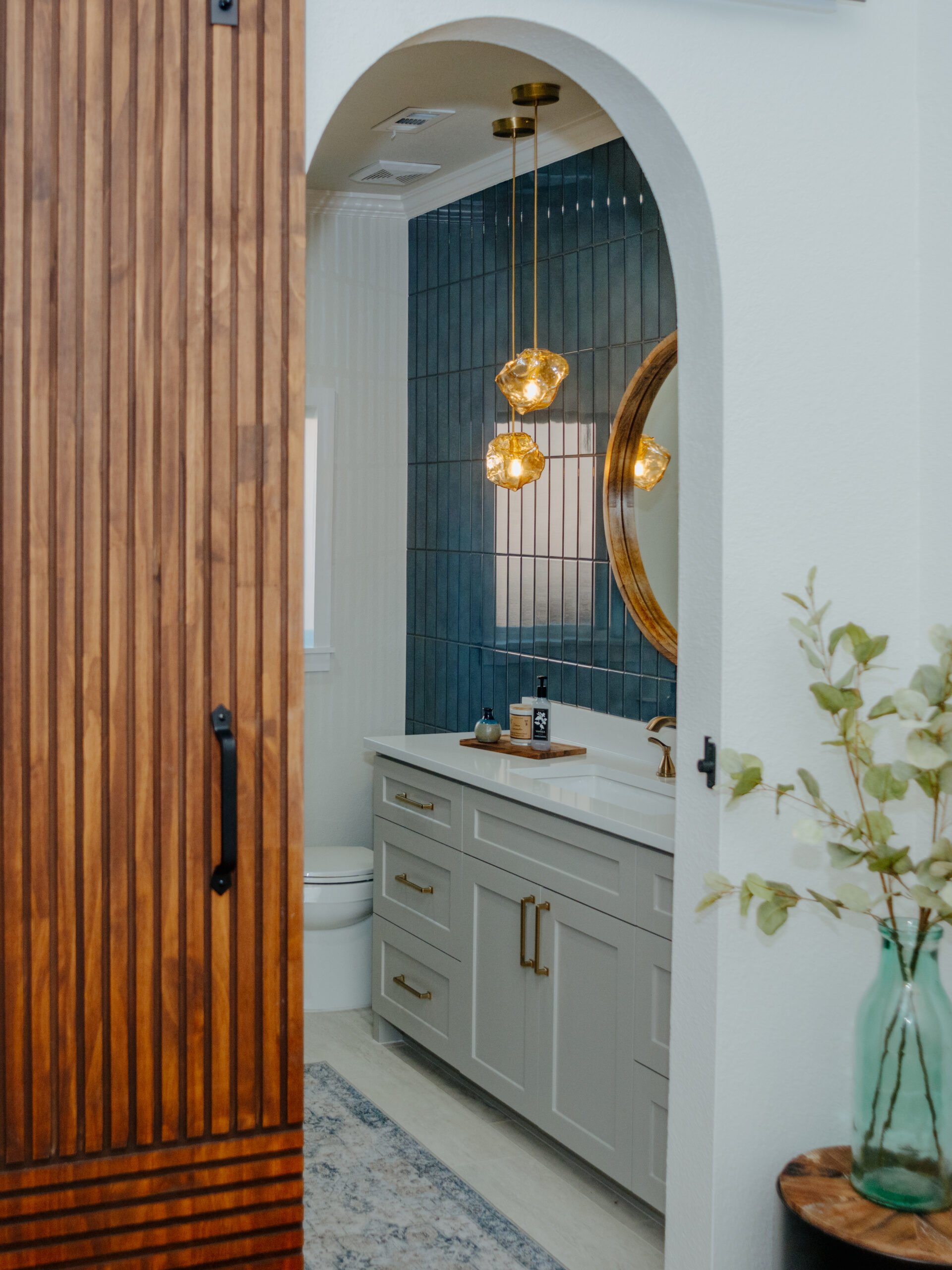 A bathroom with a white vanity, blue tiled wall, and a large round mirror. A pendant light with three glass fixtures hangs above the vanity. The door is made of vertical wooden slats, partially open, revealing the bathroom interior.