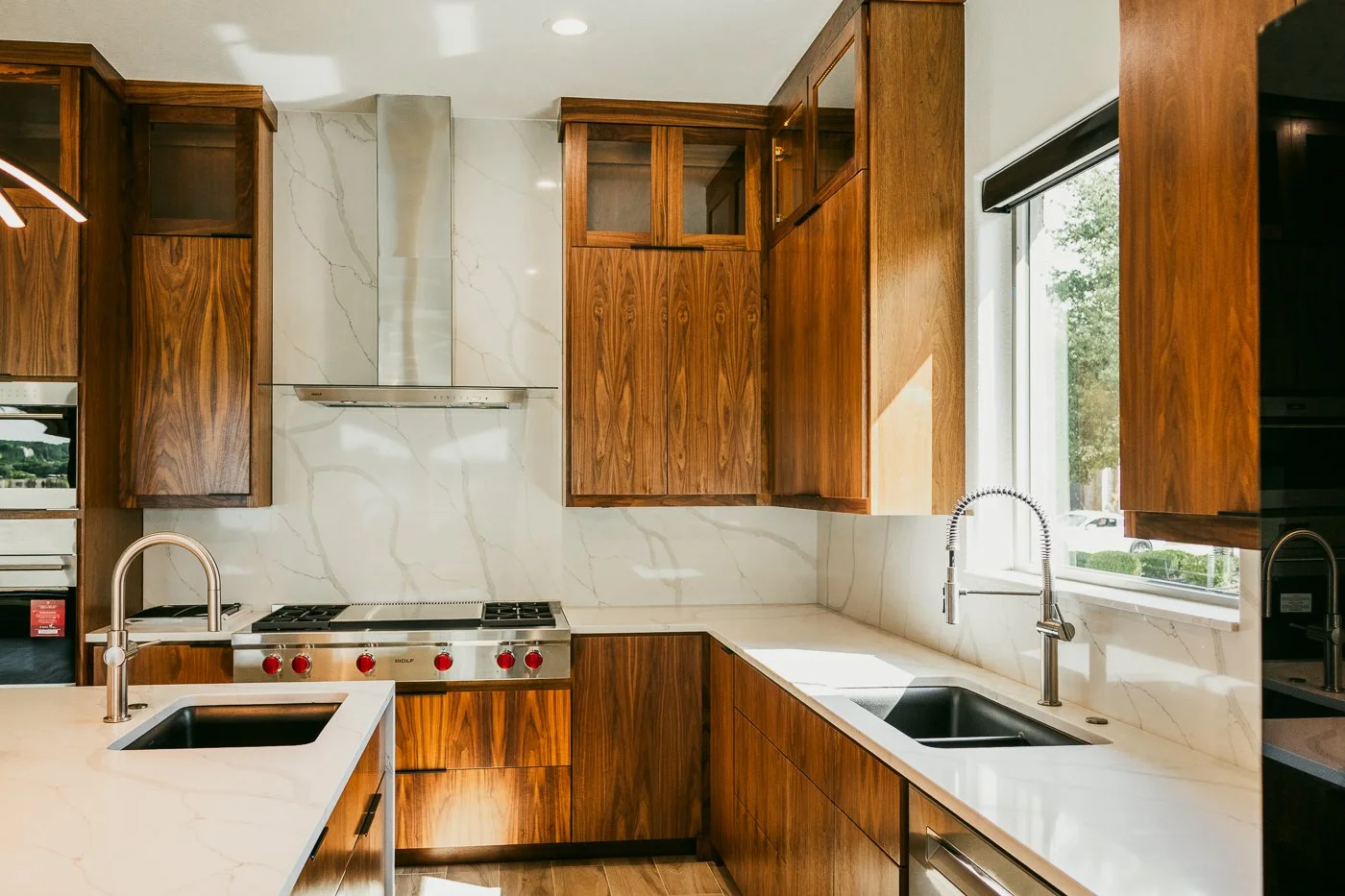 Modern kitchen with wooden cabinets, marble countertops, and two sinks, one beside a window.