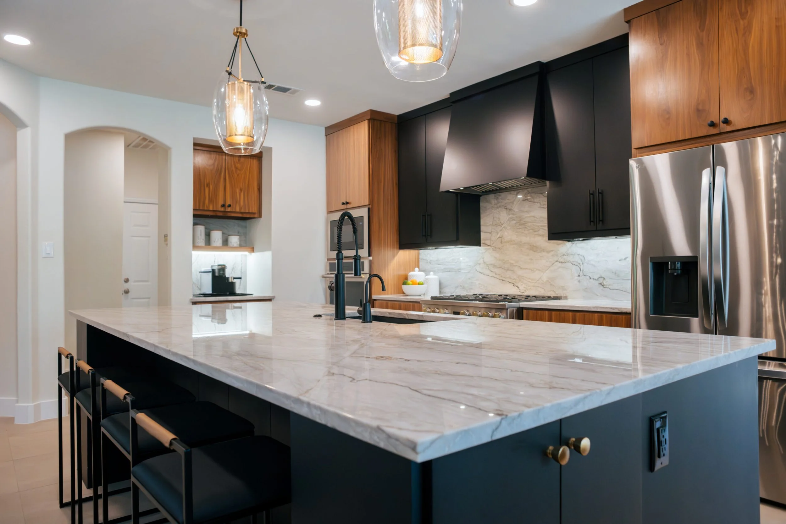 Modern kitchen with white marble island, black cabinets, wooden upper cabinets, stainless steel refrigerator, black faucet, and decorative pendant lights.