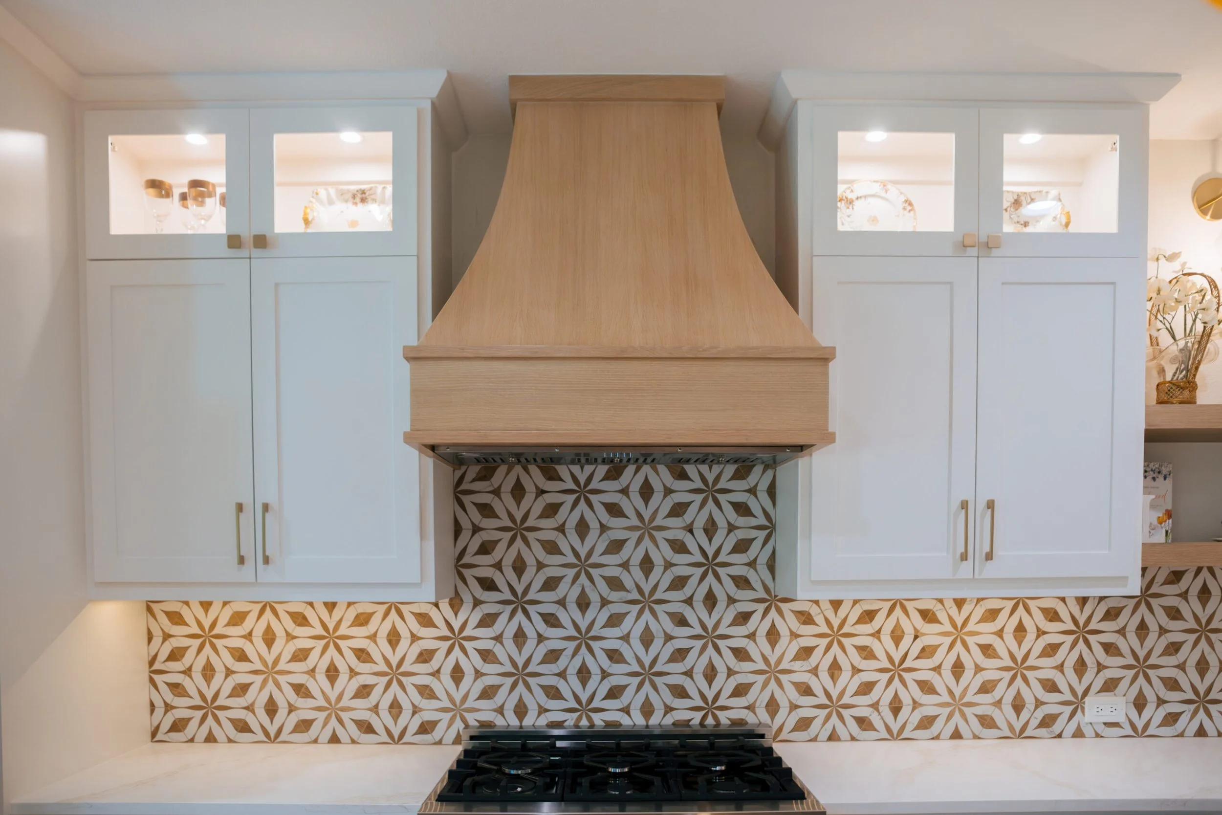 Kitchen view showing white cabinets with glass doors, a wooden range hood, patterned tile backsplash, and a gas cooktop.