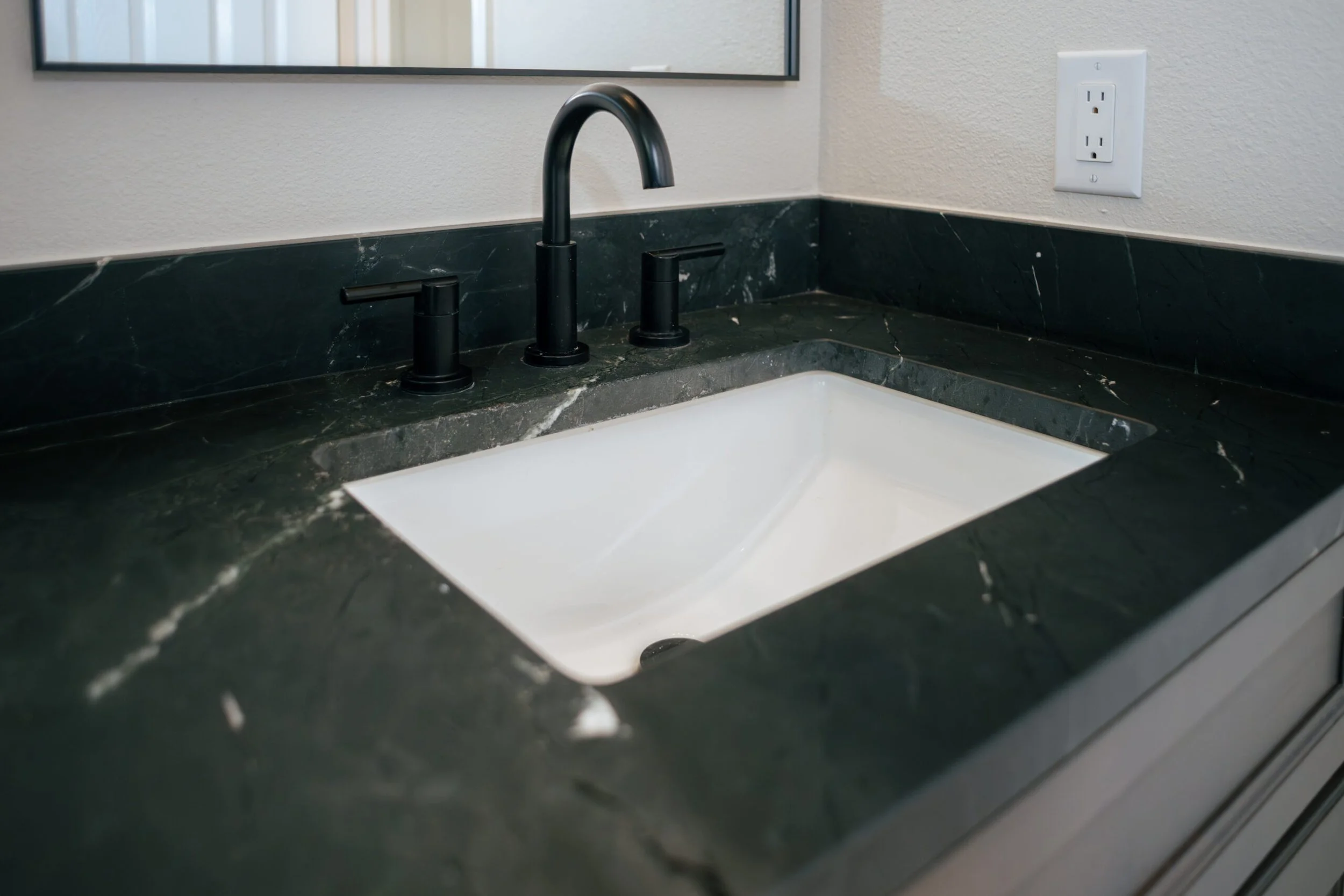 Close-up of a bathroom sink with a black faucet and a black soap dispenser on green marble countertop, reflected in a mirror. White wall with electrical outlet in background.