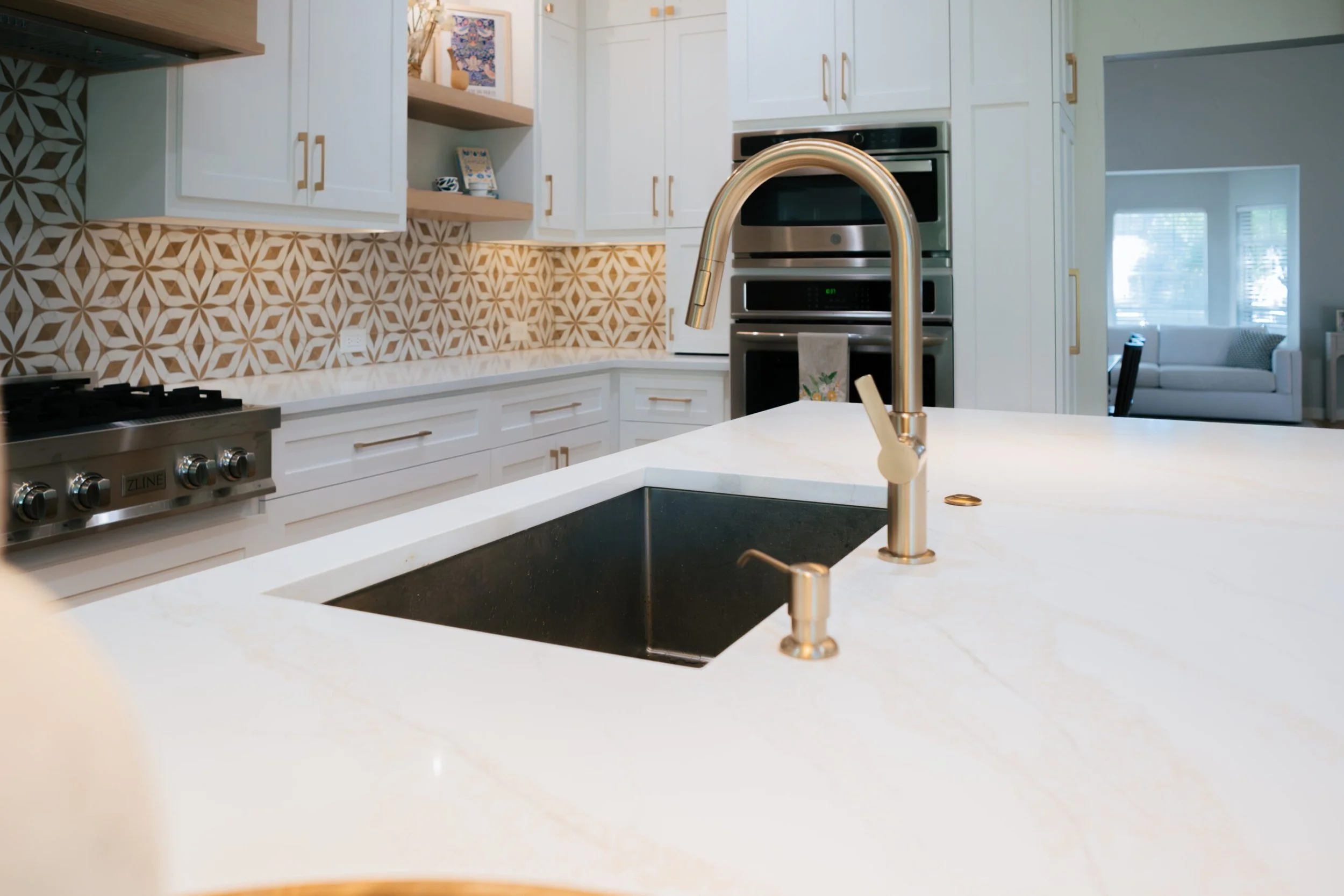 Modern kitchen with white cabinets, stainless steel appliances, a geometric backsplash, a sink with a gold faucet, and a view into a living room with a window and furniture.