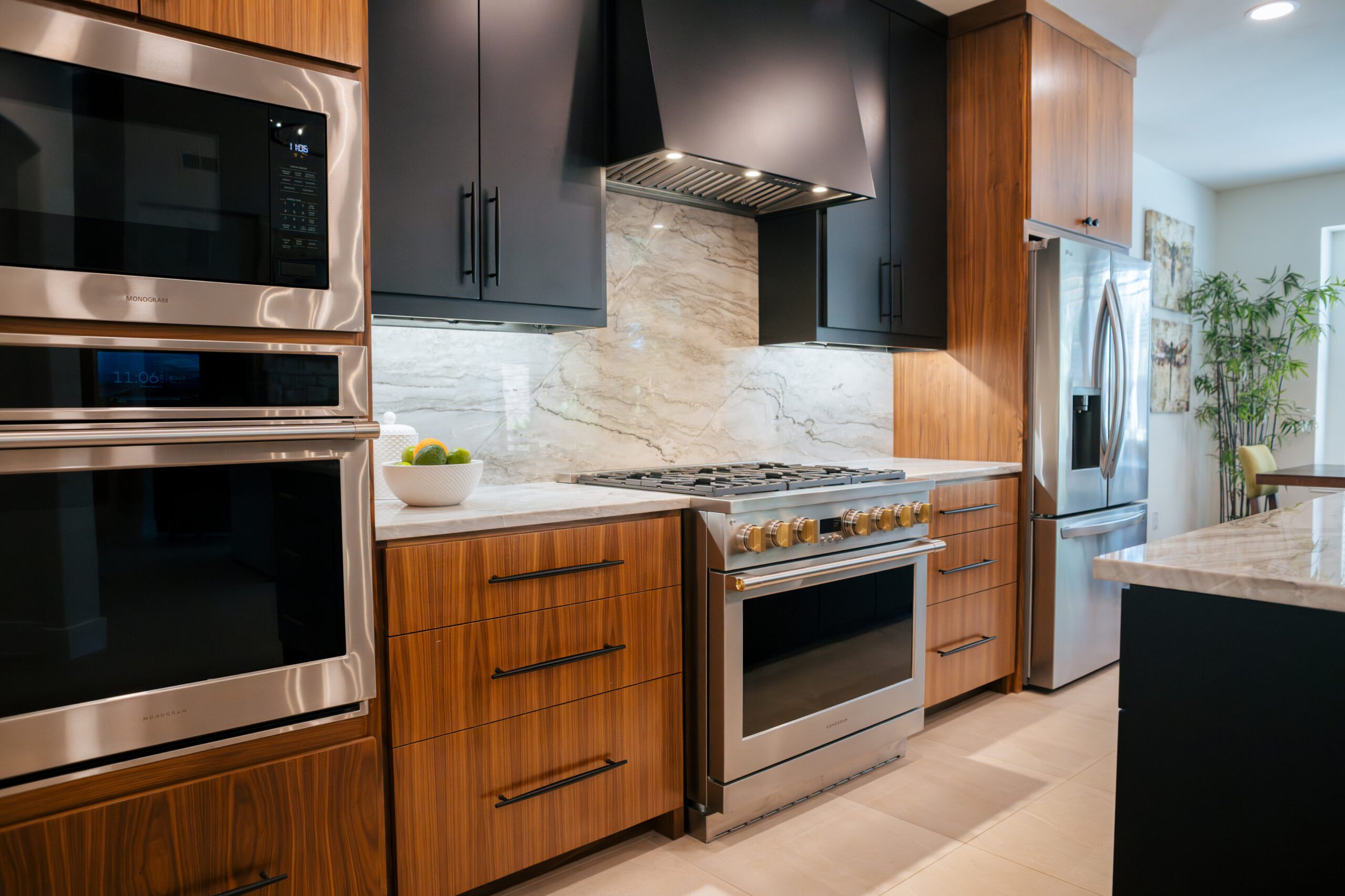Modern kitchen with black and wooden cabinets, marble backsplash, stainless steel appliances, and a bowl of fruit on the countertop.