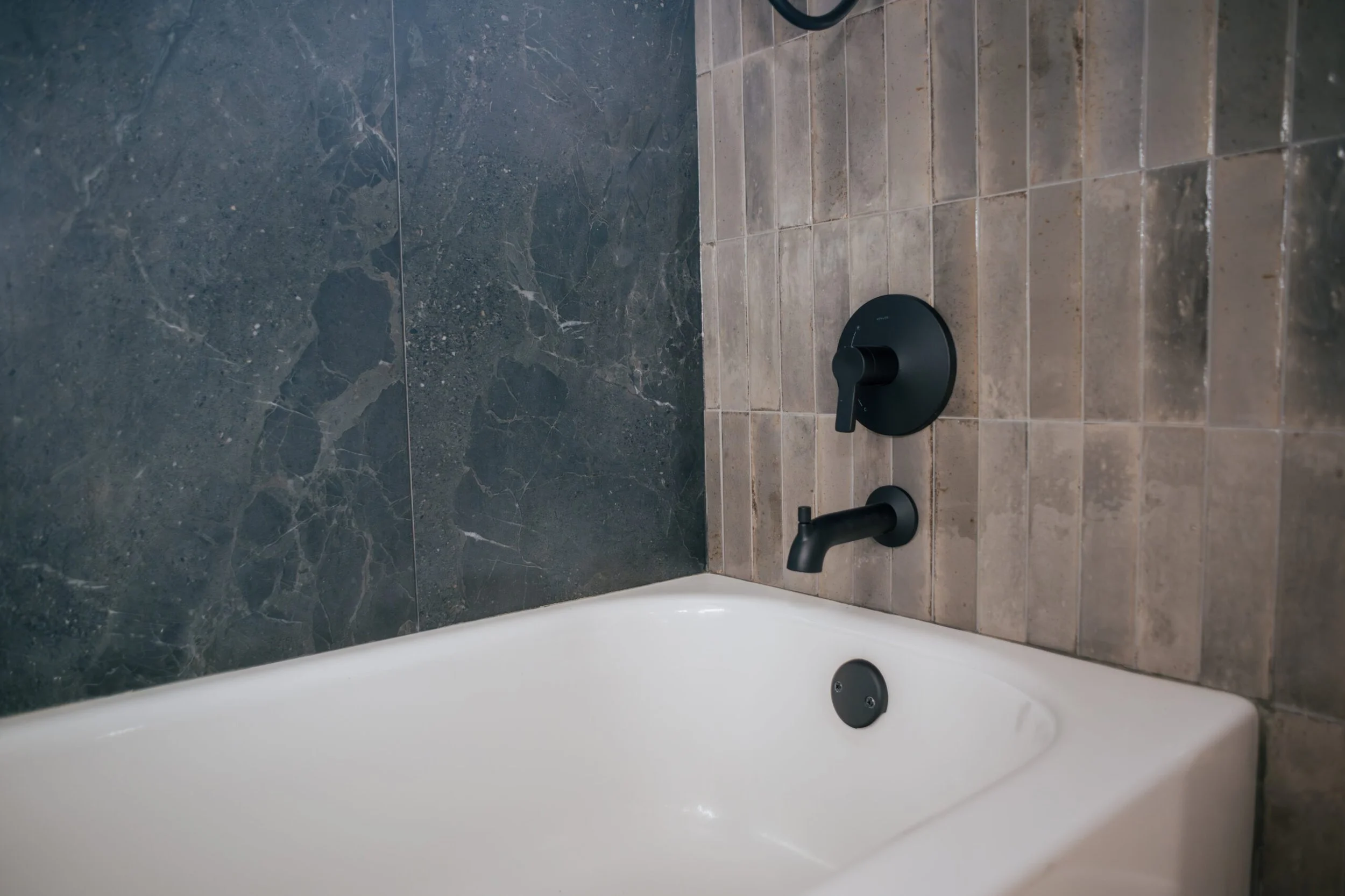 Close-up of a bathroom faucet and handles mounted on a tiled wall with dark marble and beige tiles.