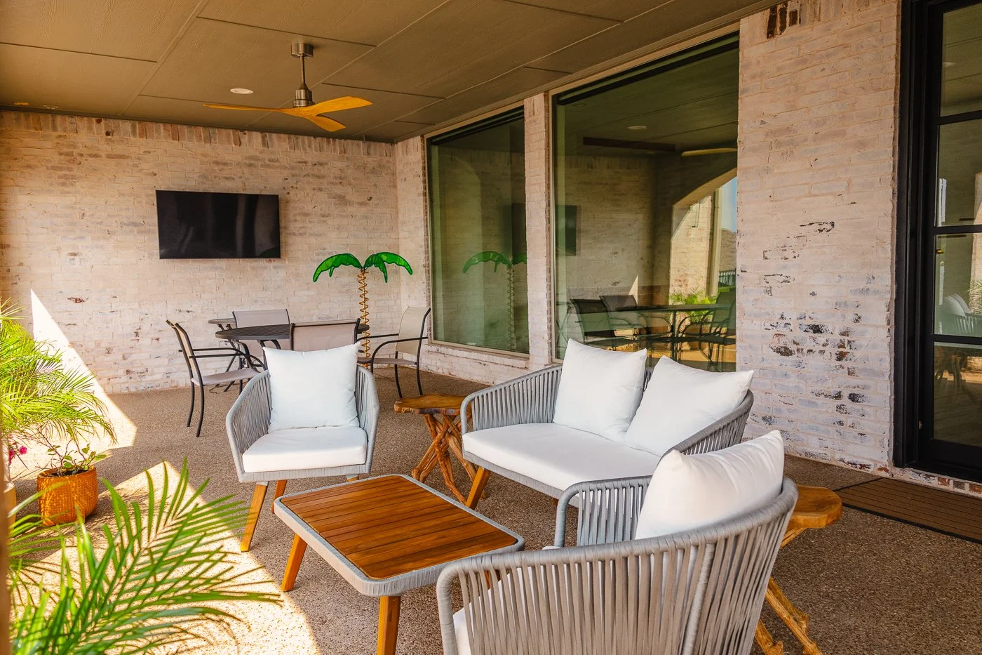 Covered outdoor patio with beige brick walls, glass sliding doors, ceiling fan, a mounted flat-screen TV, and outdoor furniture including a cushioned armchair, a cushioned loveseat, small wooden tables, and a glass-top table with chairs. Potted plant