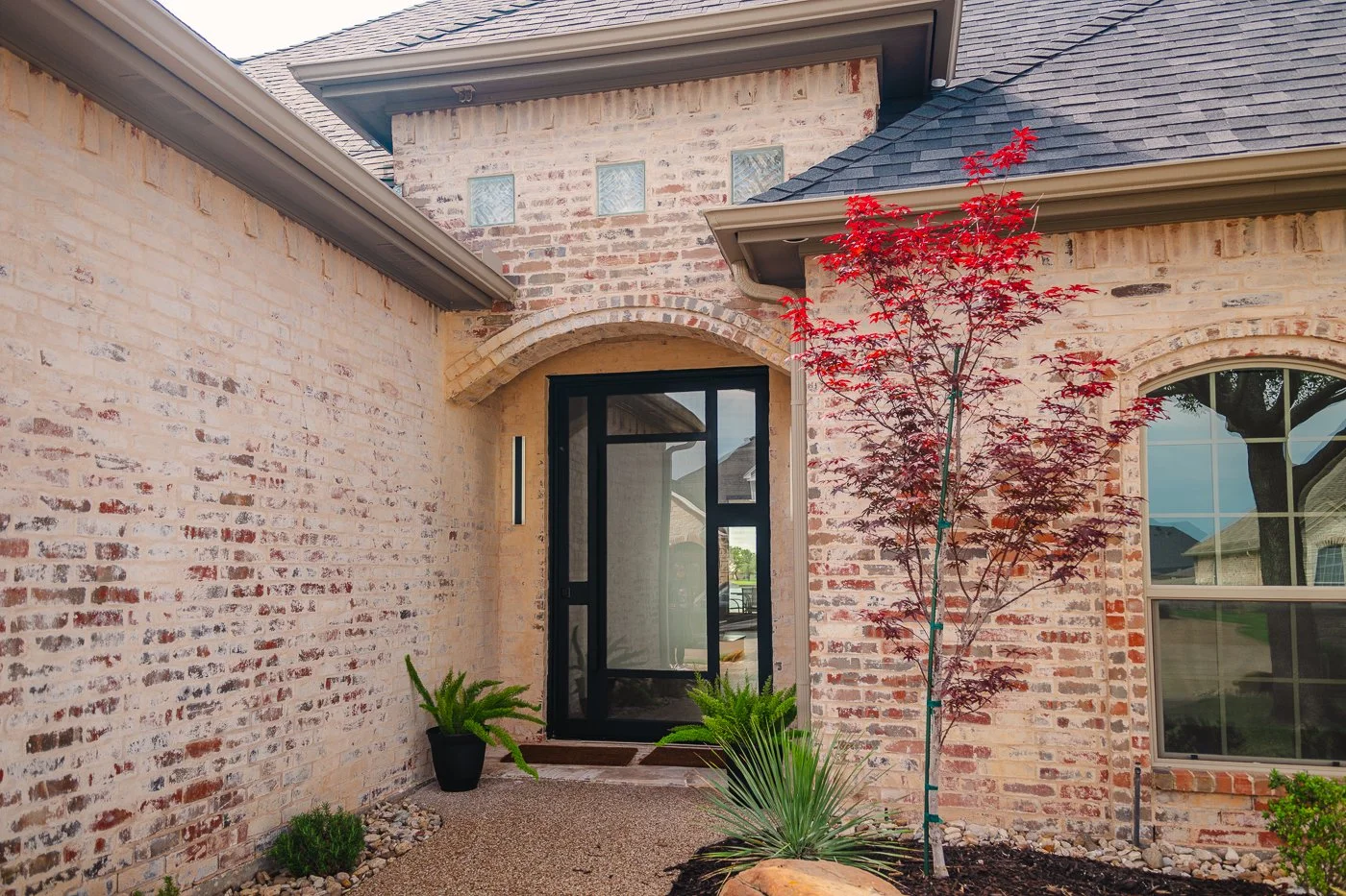 Front yard of a house with red brick exterior, black front door, small red-leafed tree, and various green plants in a landscaped garden.