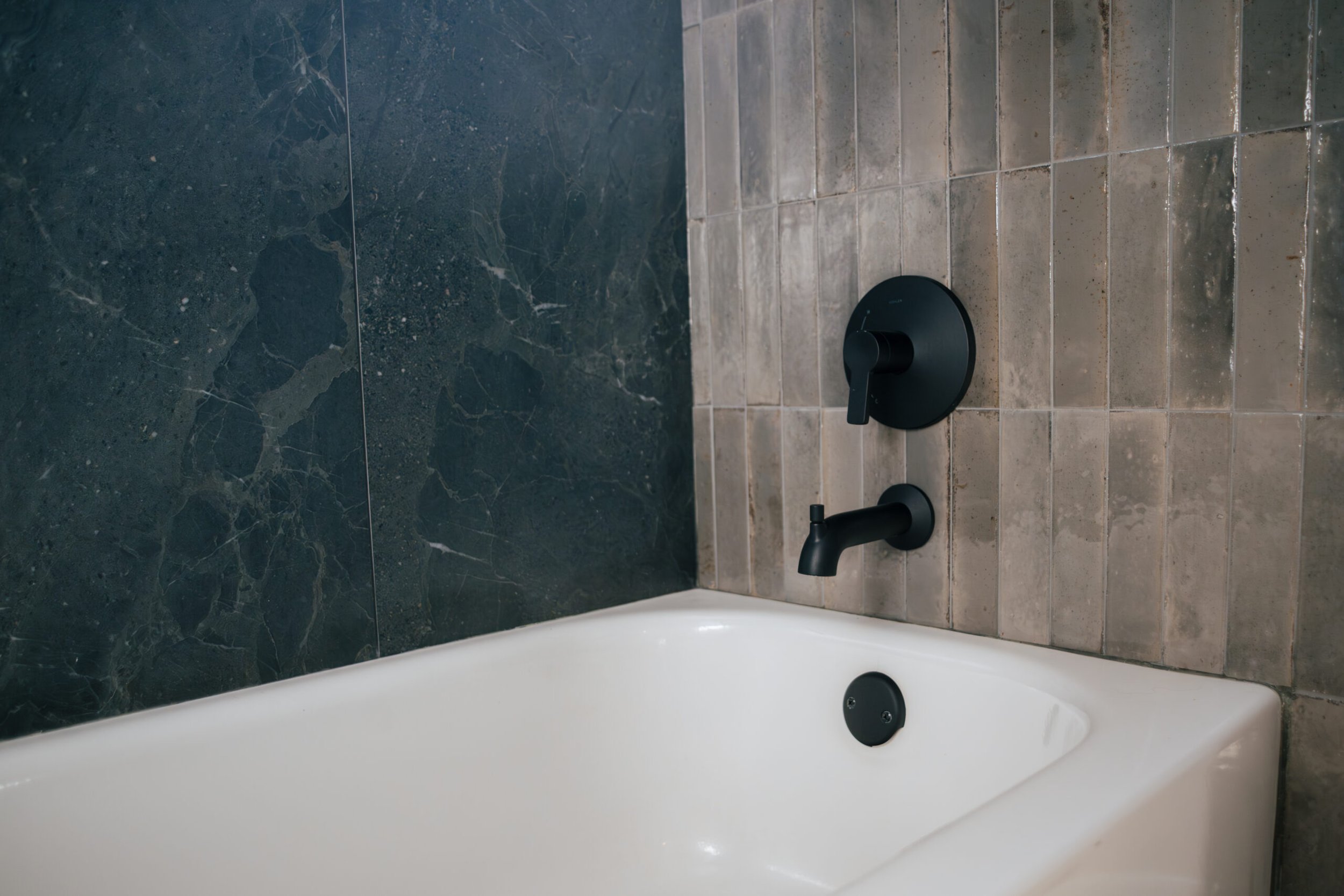 Close-up of a bathtub faucet and fixtures with a black handle, set against a tiled wall in a bathroom.