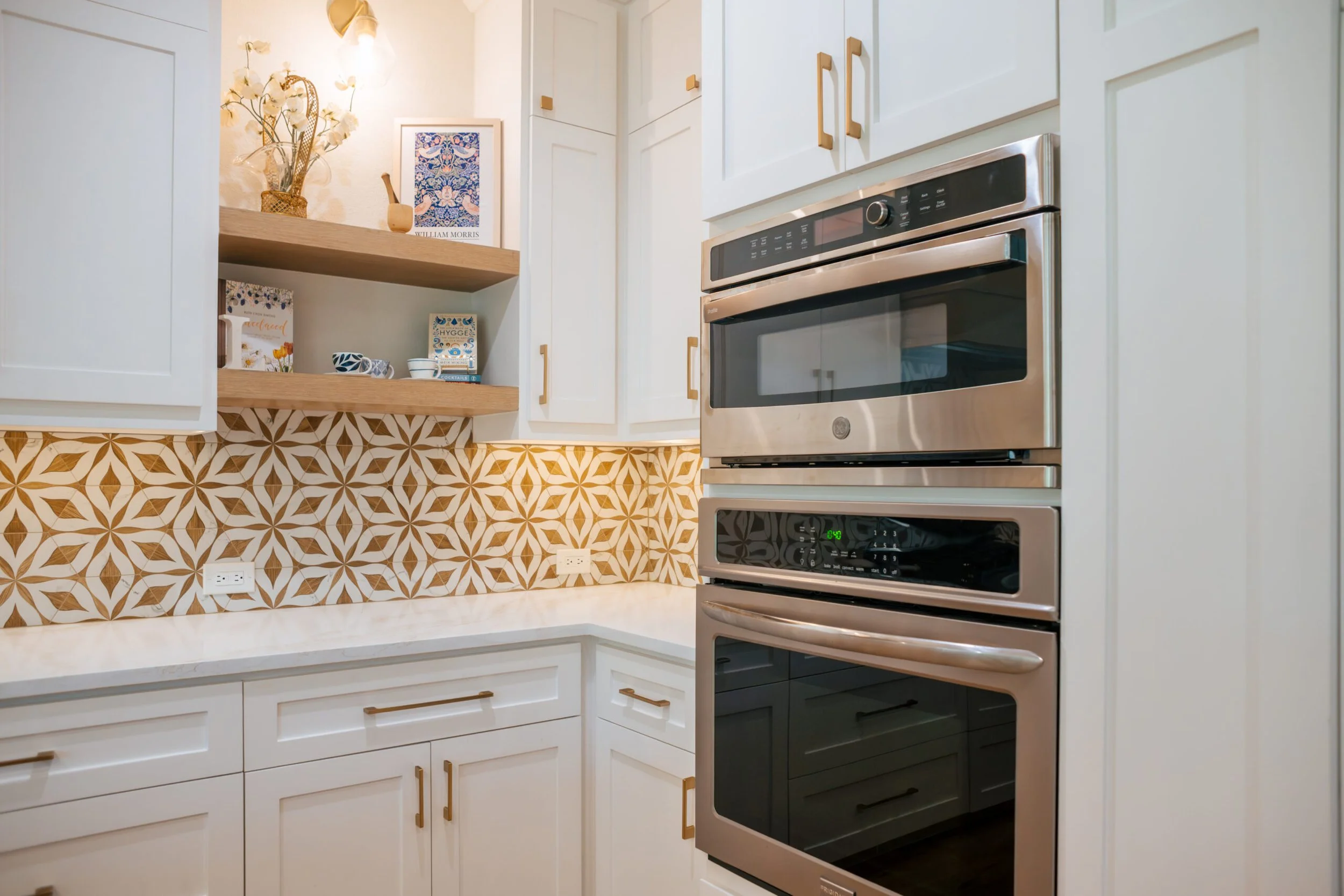 Close-up view of a modern kitchen with white cabinets, gold handles, and a stainless steel double oven. The backsplash features a brown and white geometric pattern. Open shelves hold decorative items including framed artwork, plates, bowls, and books