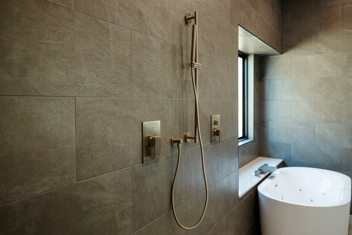 Modern bathroom with gray tiled walls, a window, and a white whirlpool bathtub. There is also a wall-mounted shower with a handheld showerhead and two controls.