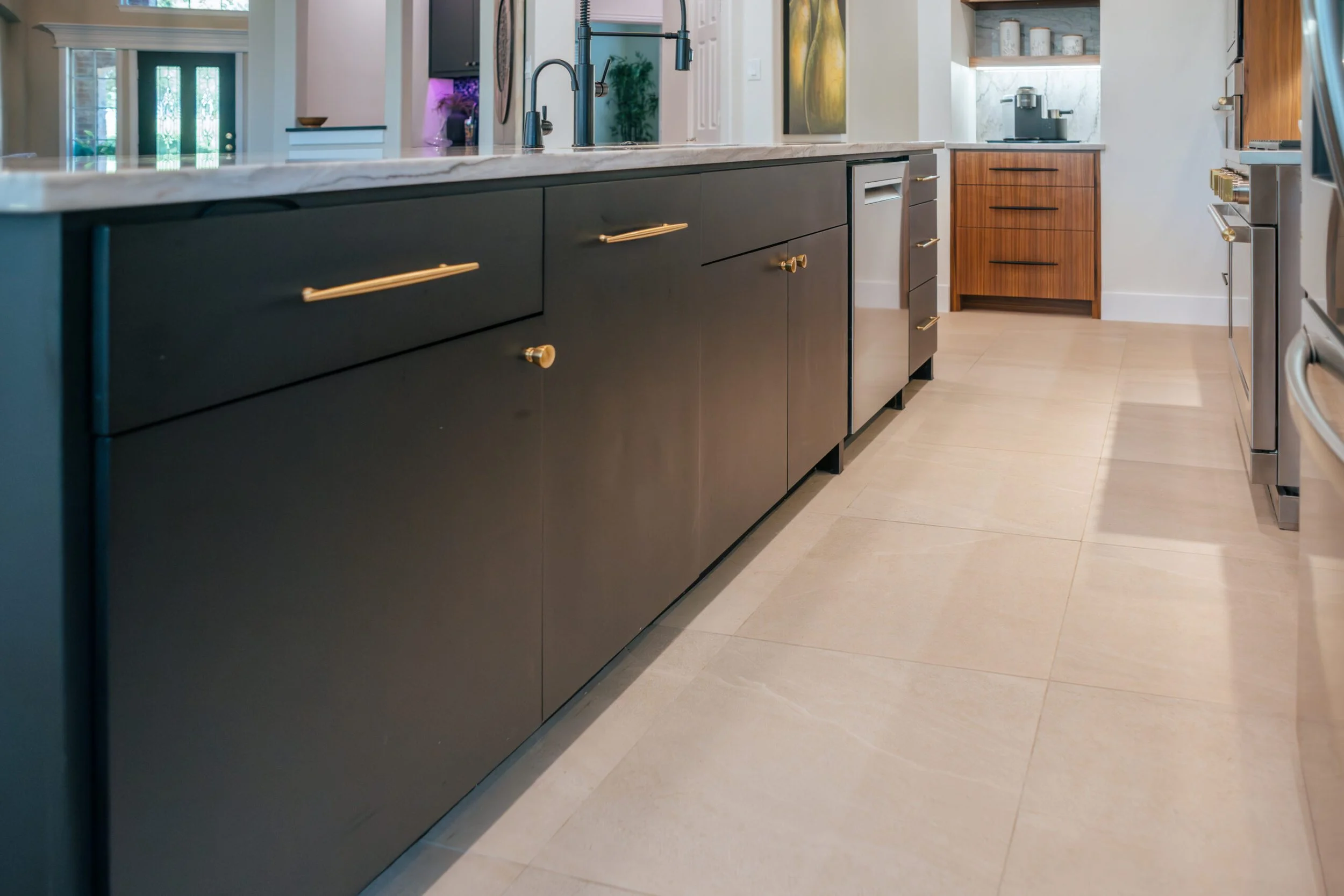Modern kitchen with dark cabinetry, gold handles, a marble countertop island, stainless steel appliances, and light wood flooring.