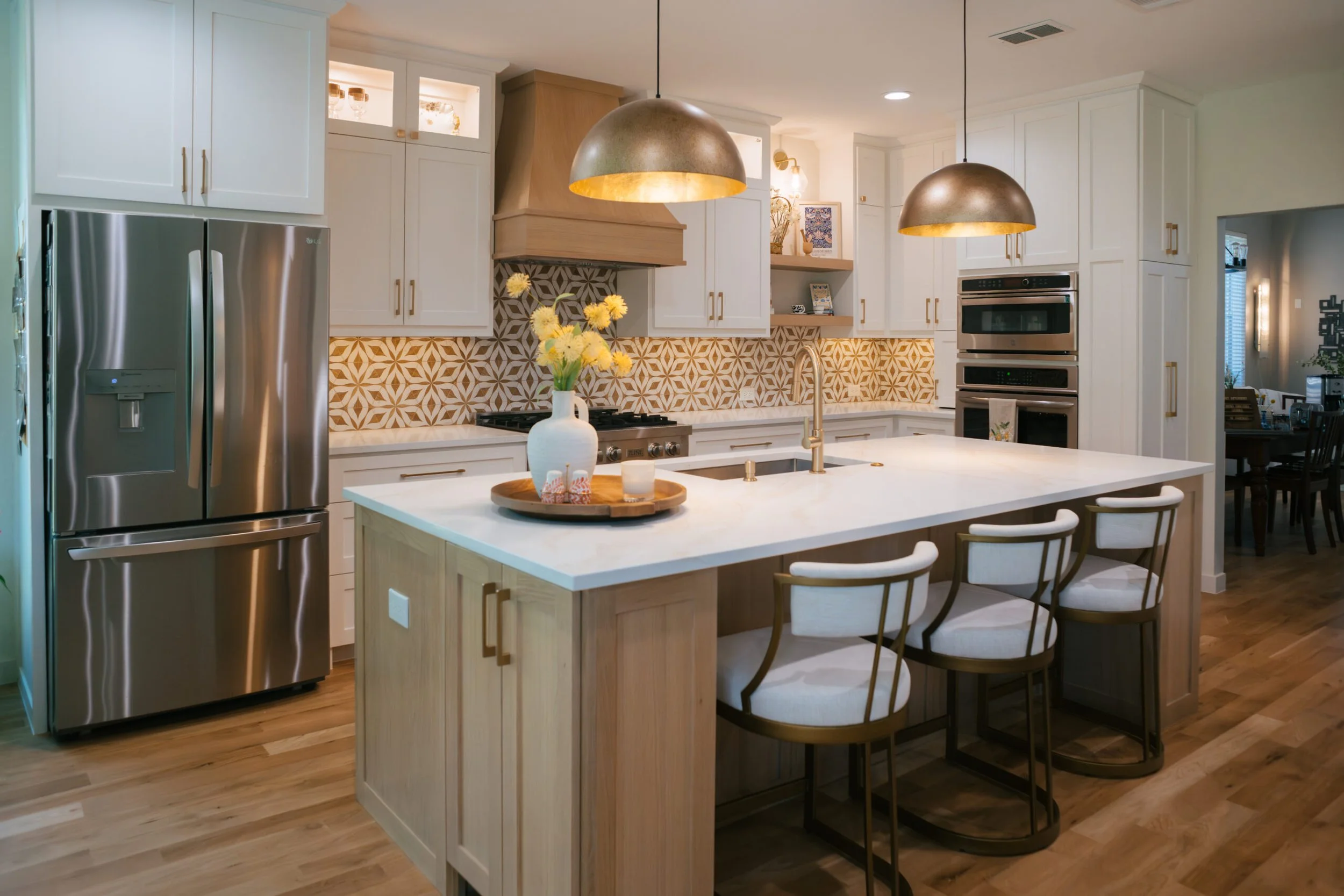 Modern kitchen with white cabinets, a patterned backsplash, a kitchen island with a vase of yellow flowers, pendant lights, stainless steel appliances, and wooden flooring.