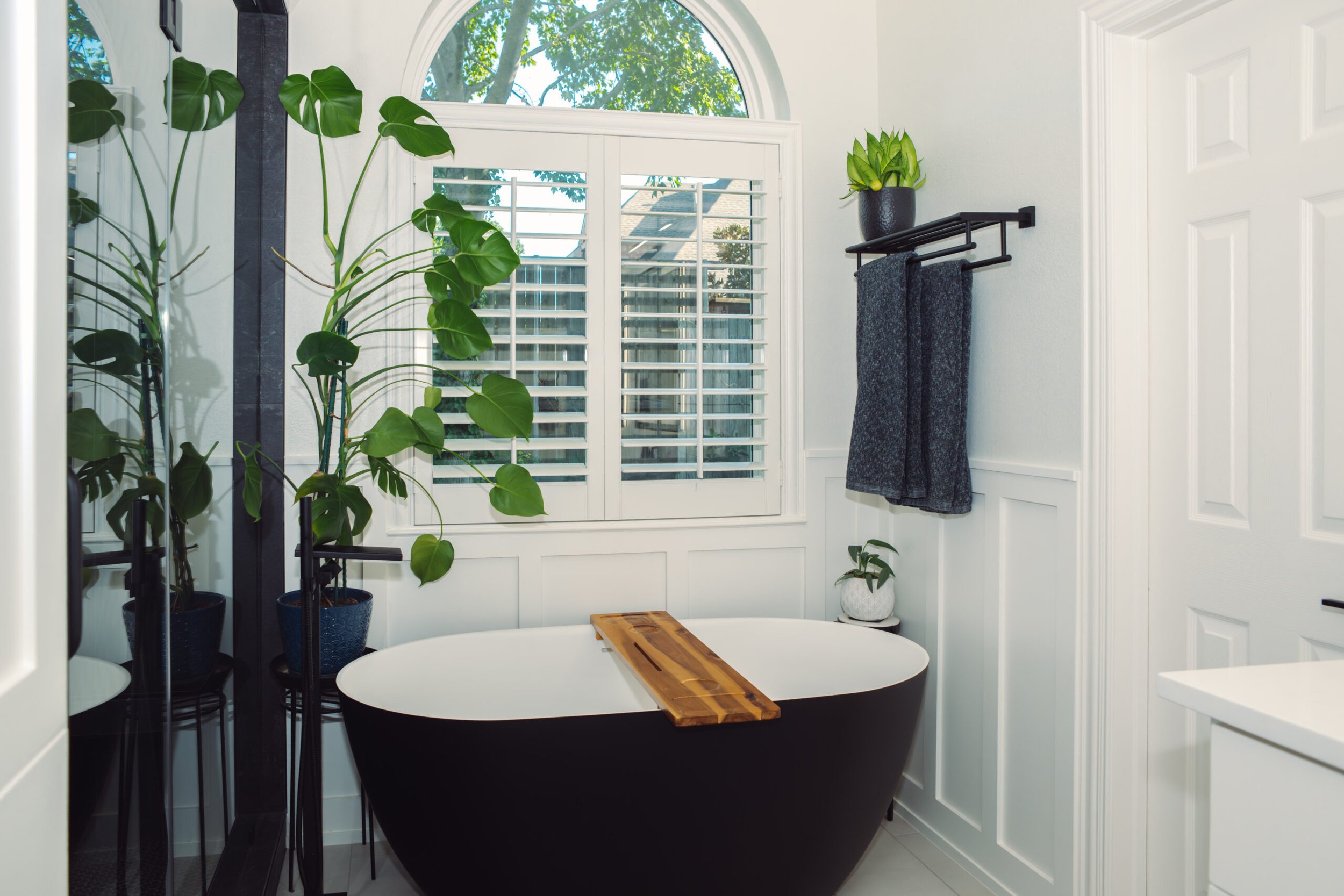 A bathroom with a black bathtub, a wooden bath tray, white walls, and a large window with white shutters. There are green houseplants near the window.