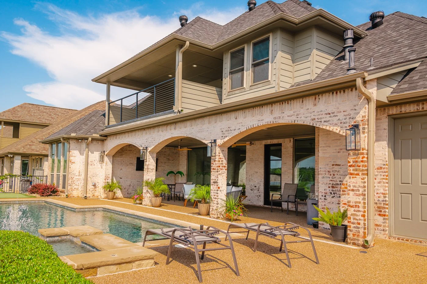Backyard of a two-story house with a swimming pool, lounge chairs, potted plants, and an outdoor seating area, featuring arched brick columns and a balcony.