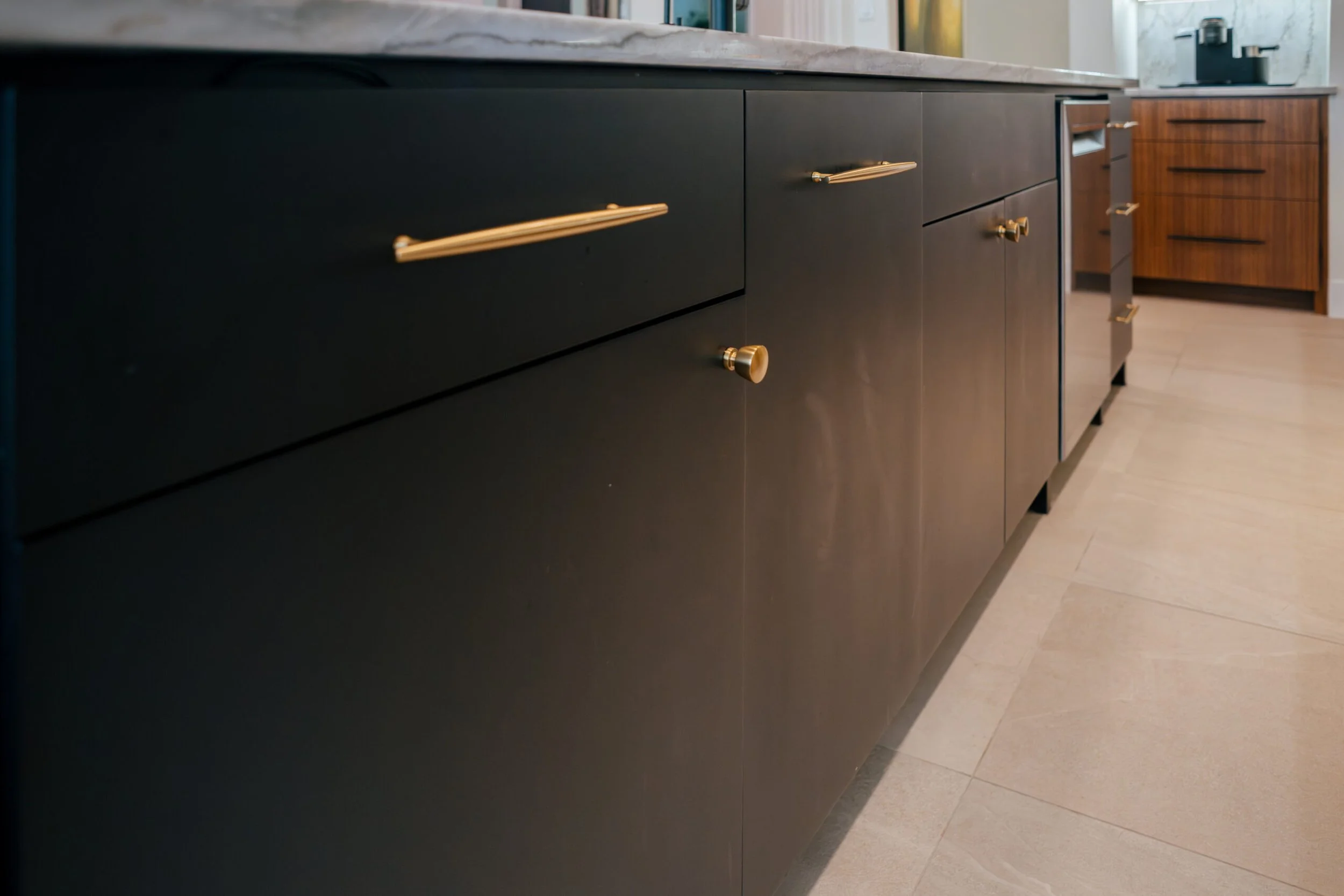Close-up of black kitchen cabinet with gold handles and knobs, with a marble countertop and wooden cabinets in the background.