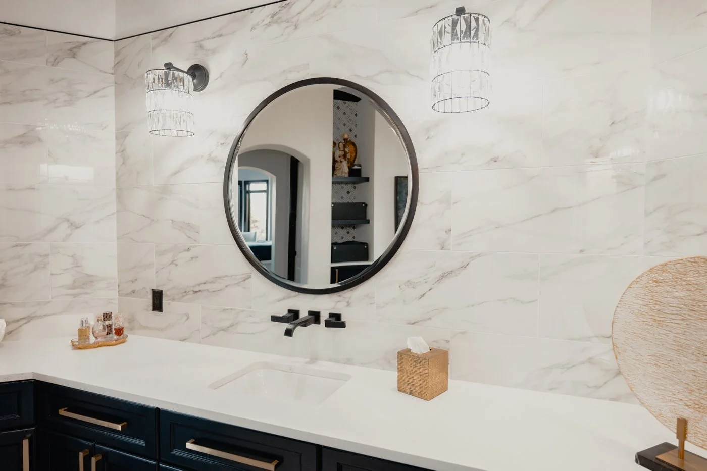 Modern bathroom with marble walls, round mirror, black vanity with gold handles, black faucet, tissue box, and decorative lighting.