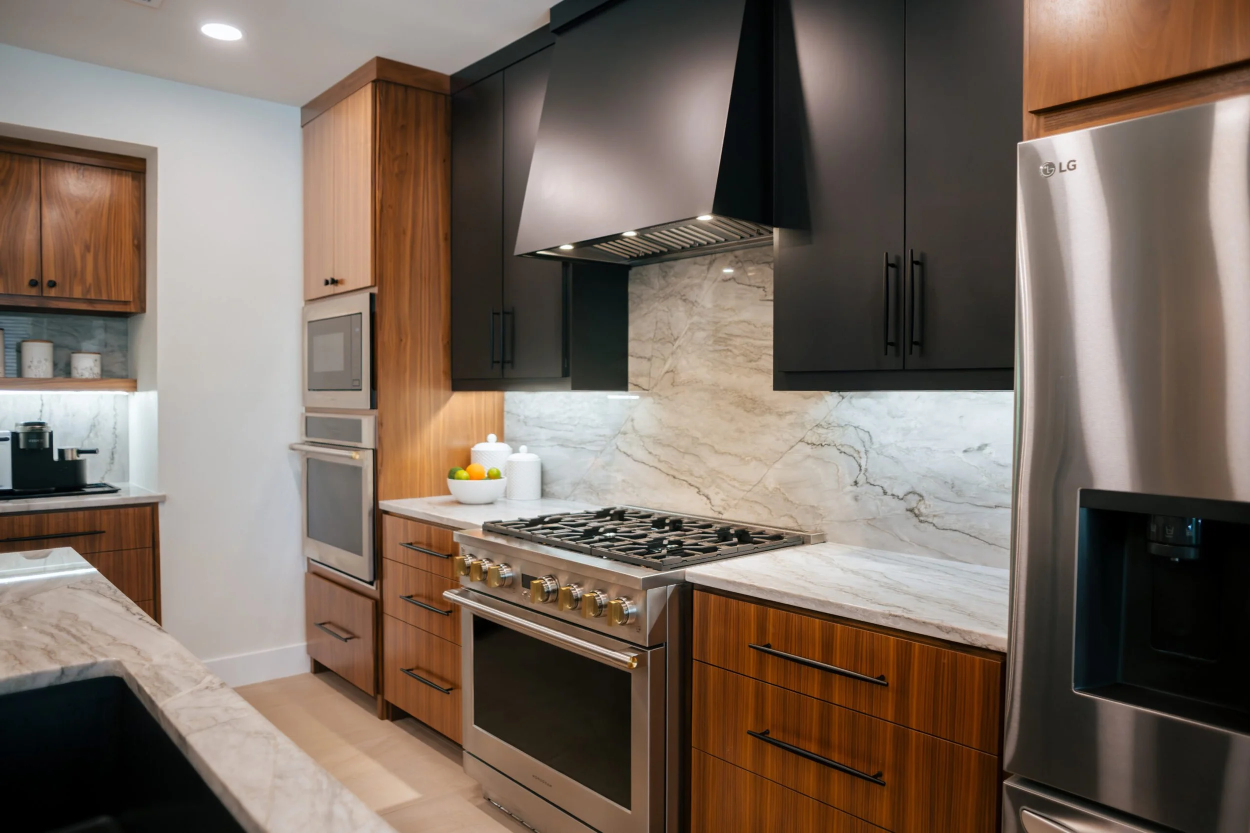 Modern kitchen with wooden and black cabinets, stainless steel oven and refrigerator, marble countertops and backsplash, and a bowl of fruit on the counter.
