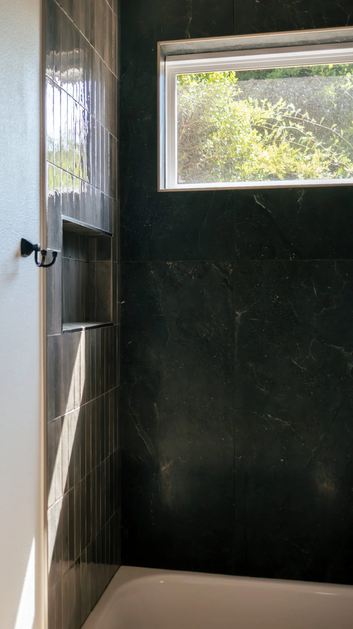 Inside view of a shower with black marble tiles, a built-in niche, and a window showing green trees outside.