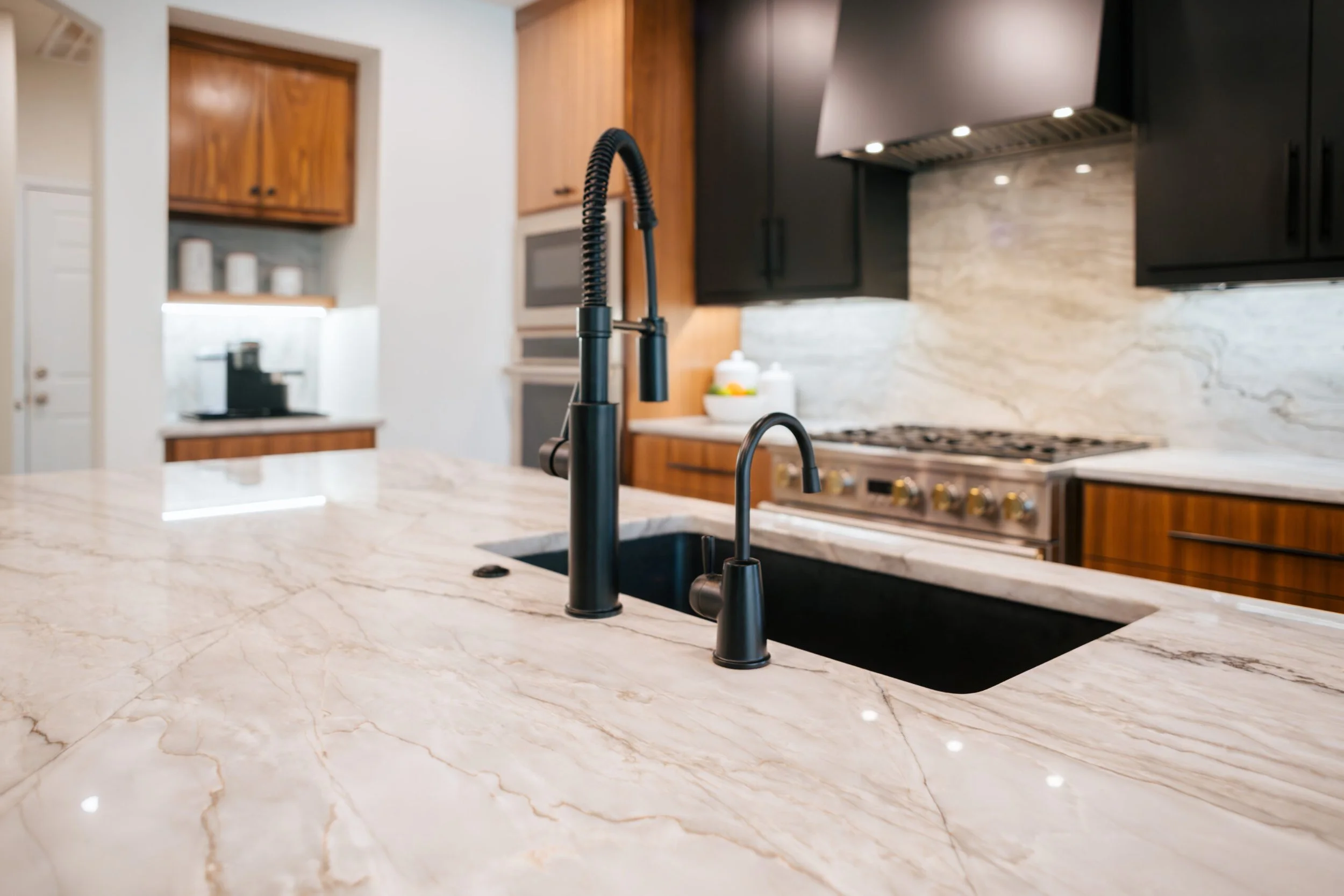 A modern kitchen with a marble countertop, black sink, black faucet, wooden cabinets, stainless steel range, and a backsplash with natural stone pattern.