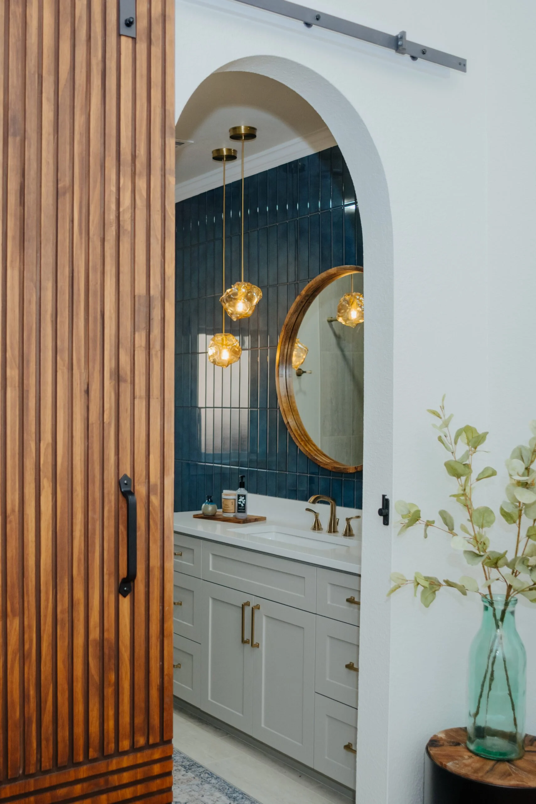 Interior of a modern bathroom with a white vanity, a round wooden framed mirror, blue tiled wall, brass fixtures, and decorative pendant lights. Part of a wooden sliding door is visible in the foreground, and a large glass vase with green leaves is on a small table.
